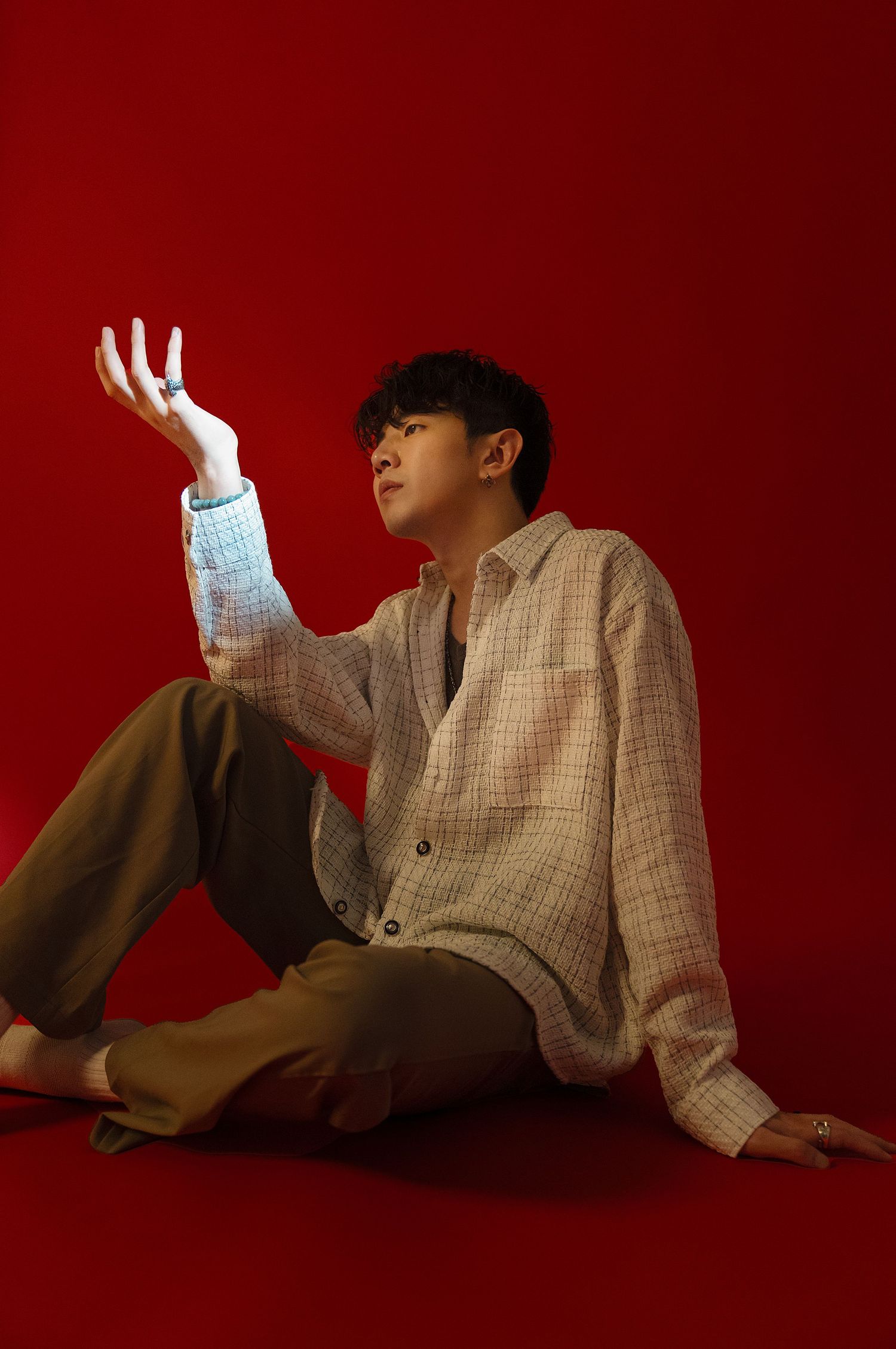 Young man in plaid shirt sitting on red background, reaching hand upward in artistic pose.