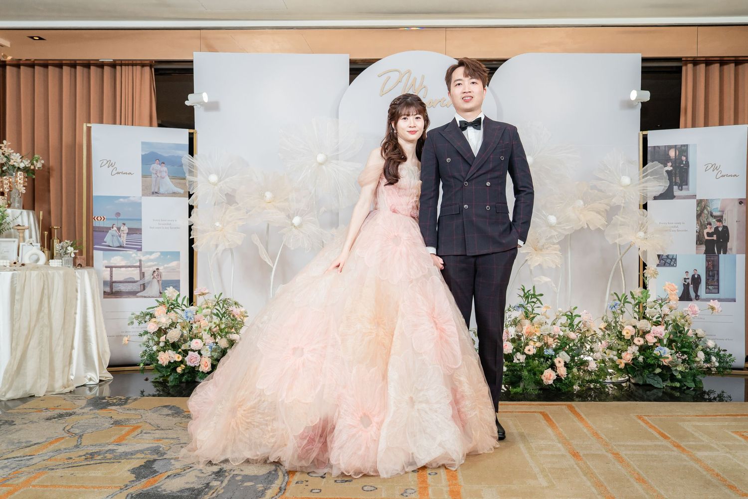 Bride in pink ballgown and groom in navy suit pose at elegant floral wedding reception backdrop.