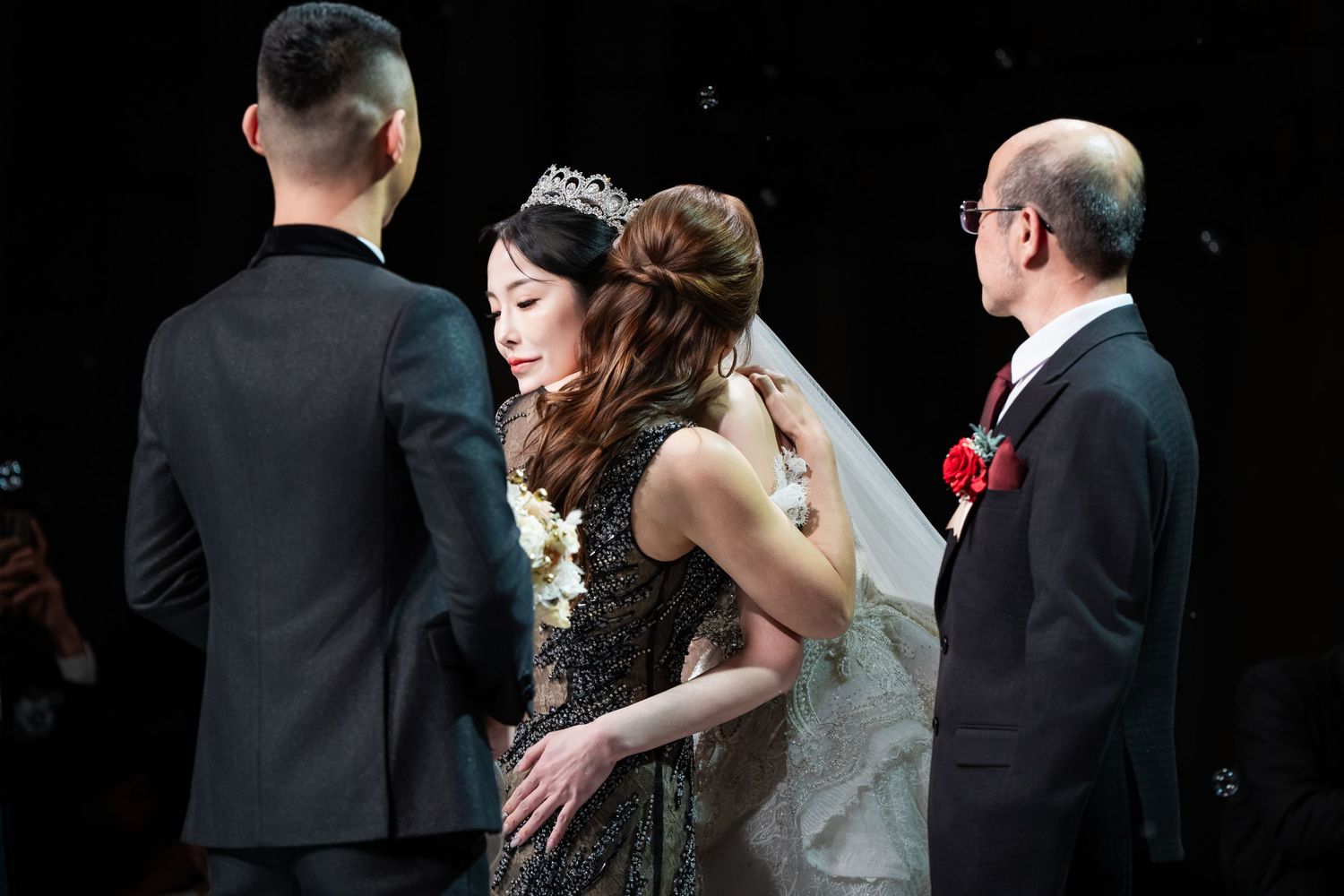 A wedding ceremony in progress with people in formal attire including a black dress and white veil against dark background.