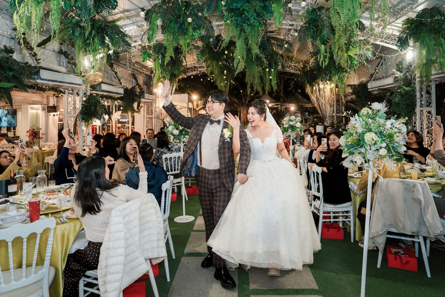 A wedding celebration in a lush indoor garden venue with elegant table settings and hanging greenery decorations.