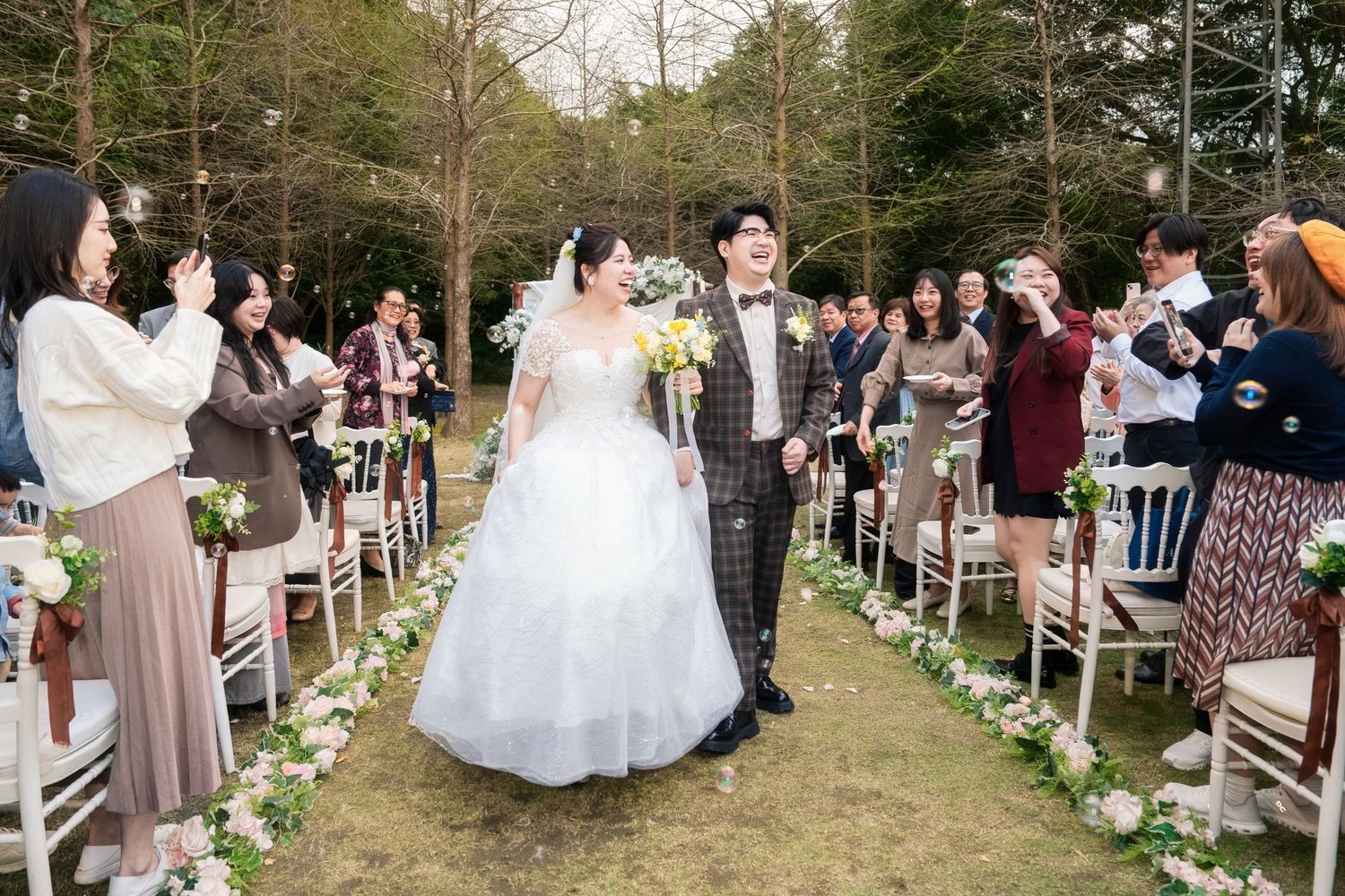 Wedding ceremony guests seated in white chairs along an outdoor aisle decorated with floral arrangements and greenery.