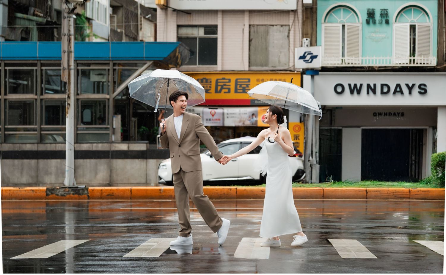 A couple holding umbrellas dances joyfully on a rainy street in front of pastel-colored storefronts.
