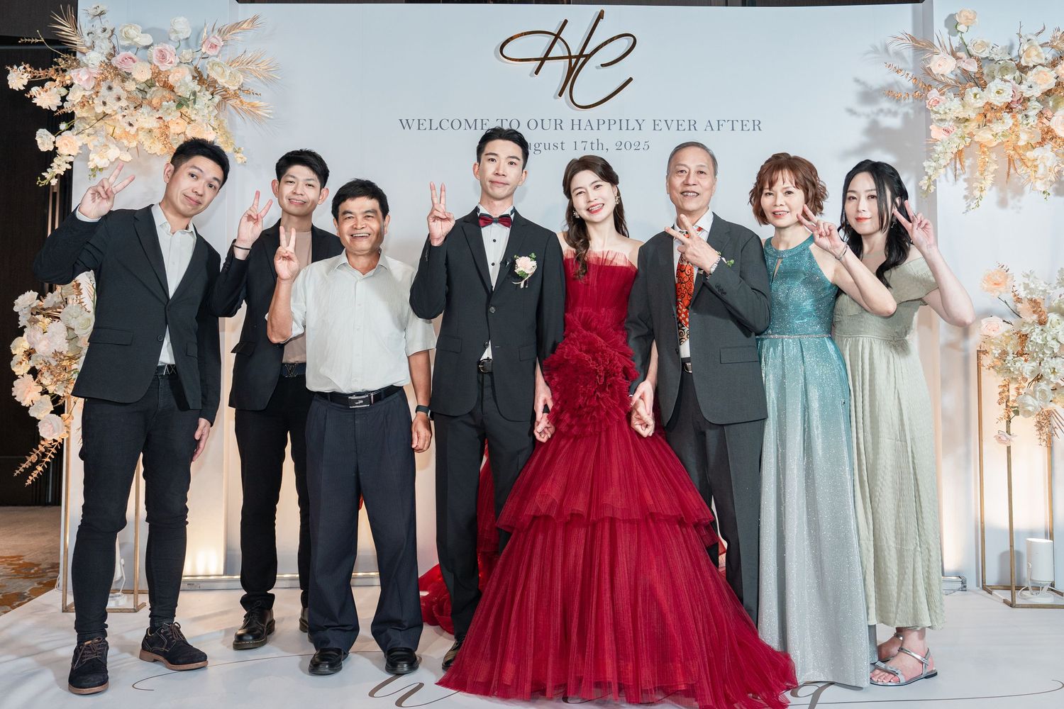 Wedding party in formal attire poses in front of decorative floral arch backdrop with bride in stunning deep red gown.