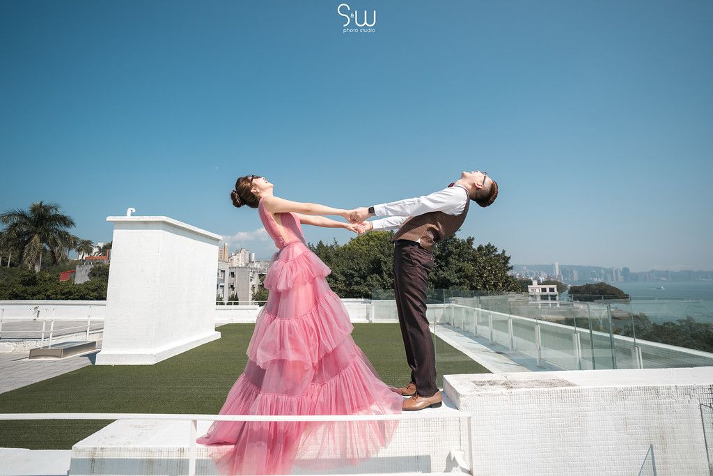 A couple poses playfully on a rooftop balcony with the person in pink leaning back dramatically against the ocean view.
