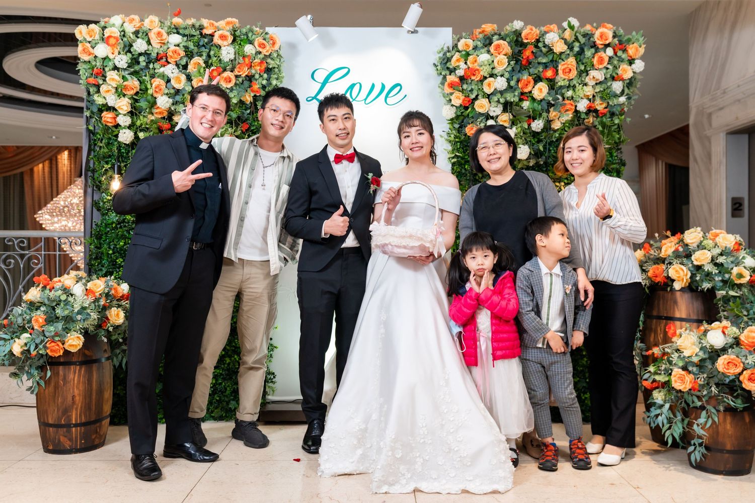 Wedding group of various ages gathers for photo in front of decorative floral backdrop.