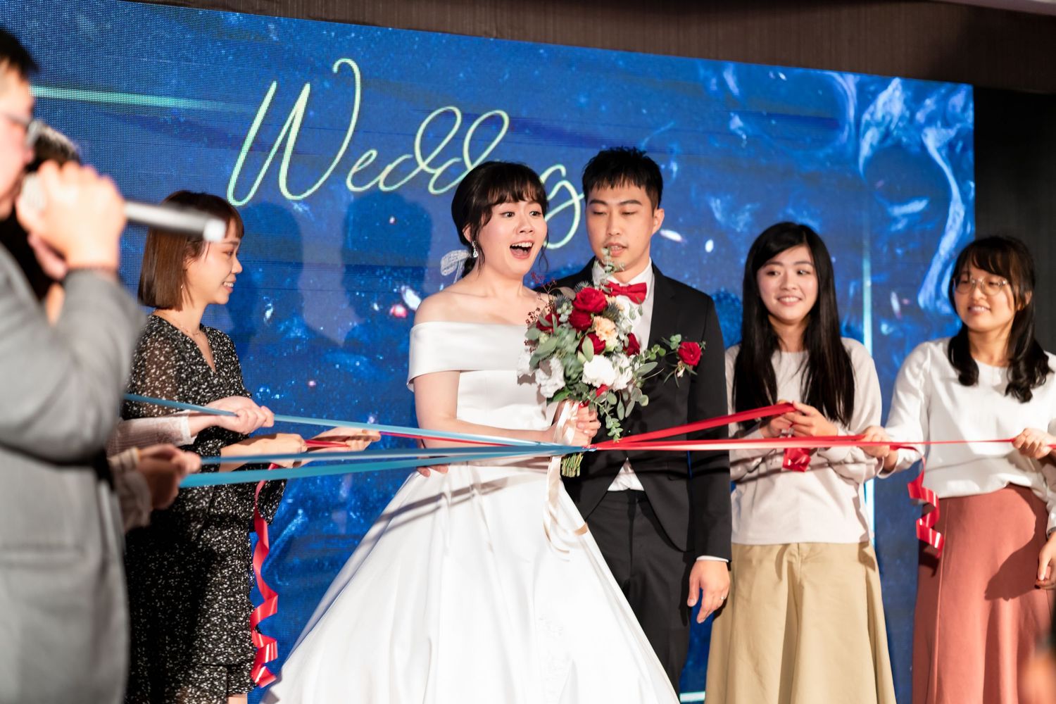 Wedding guests hold a red ribbon while celebrating with the newlyweds at a reception with a blue backdrop.