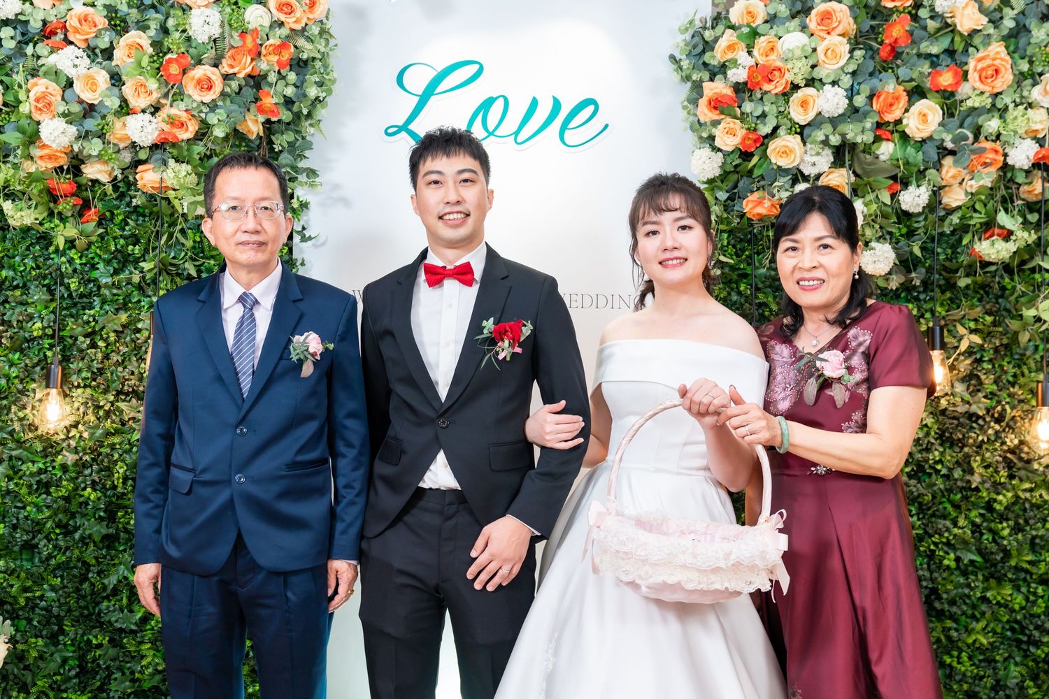 Wedding party poses in front of floral wall with turquoise 'Love' sign at elegant celebration.
