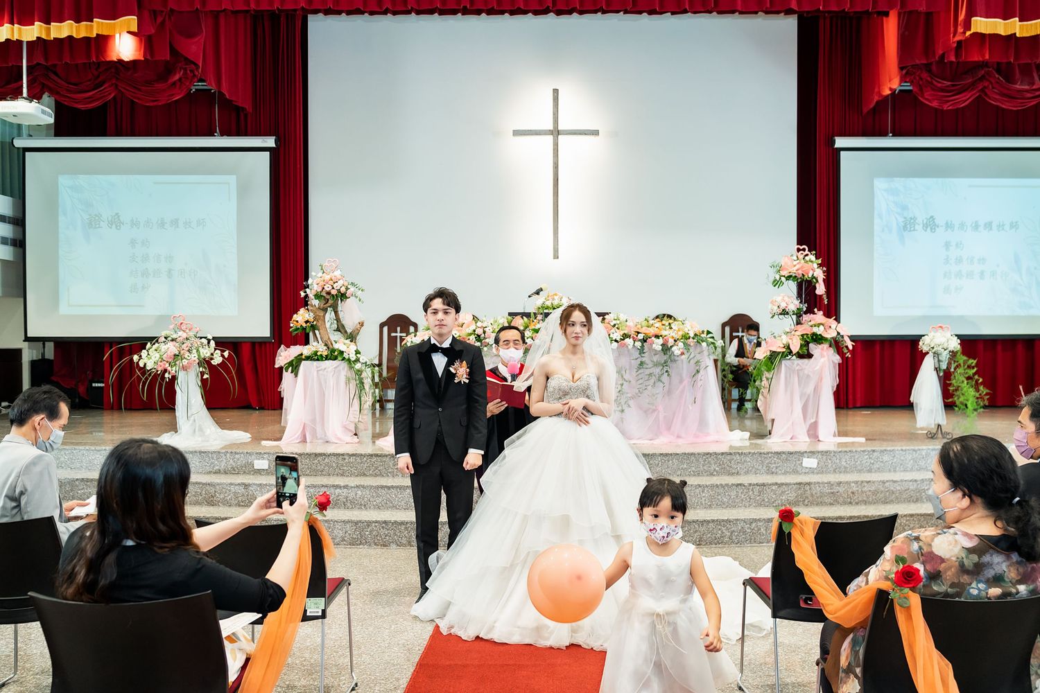 Church wedding ceremony with a large cross and red carpet aisle decorated with white floral arrangements.