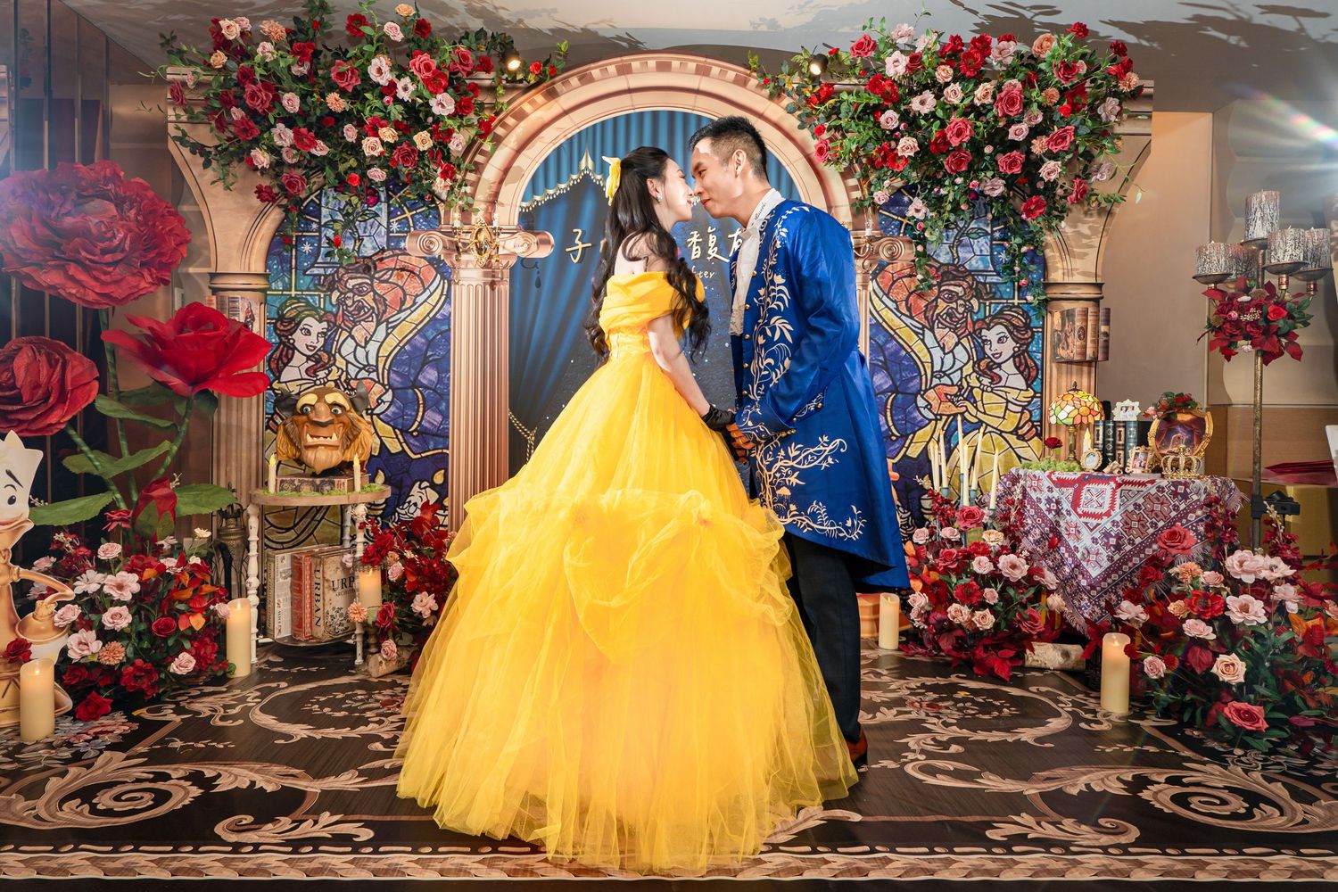 Couple in Beauty and the Beast costumes pose before floral arch backdrop at themed wedding celebration.