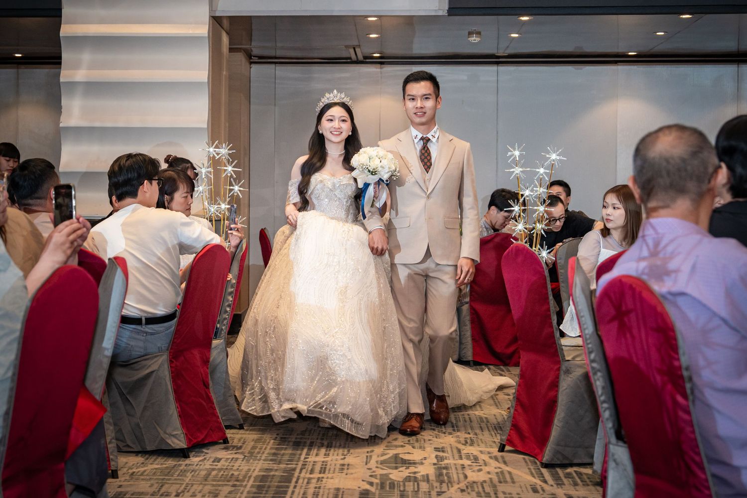Bride in white gown and groom in tan suit walk down aisle at elegant indoor wedding ceremony.
