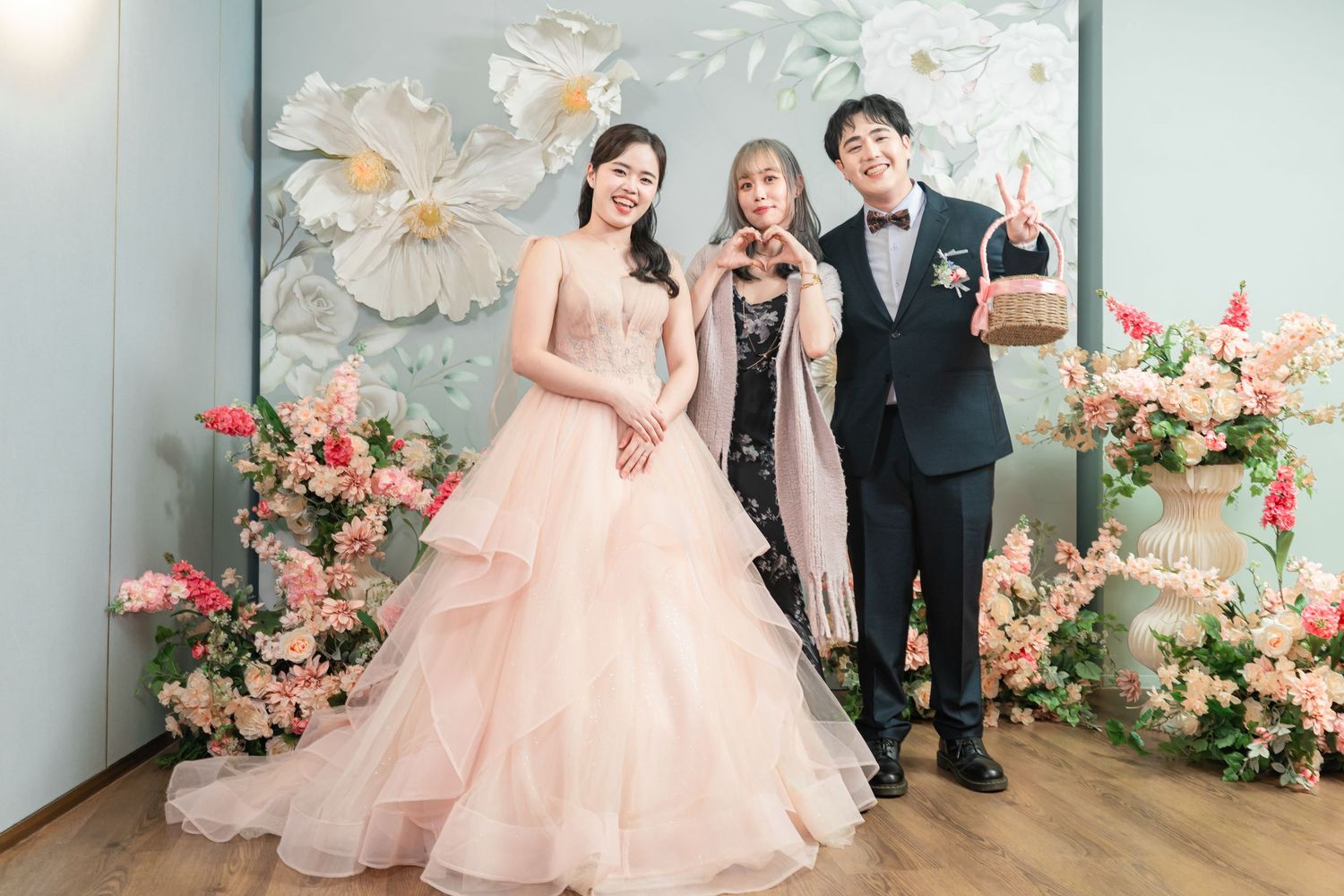 A wedding celebration scene with a pink floral backdrop and elegant formal attire against a light blue wall.