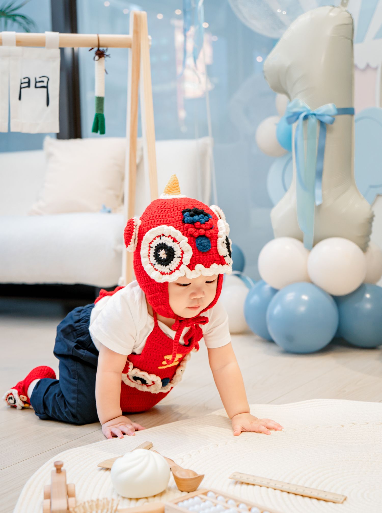 Baby in red and white holiday outfit crawls across white floor with blue and white balloon decorations in background.