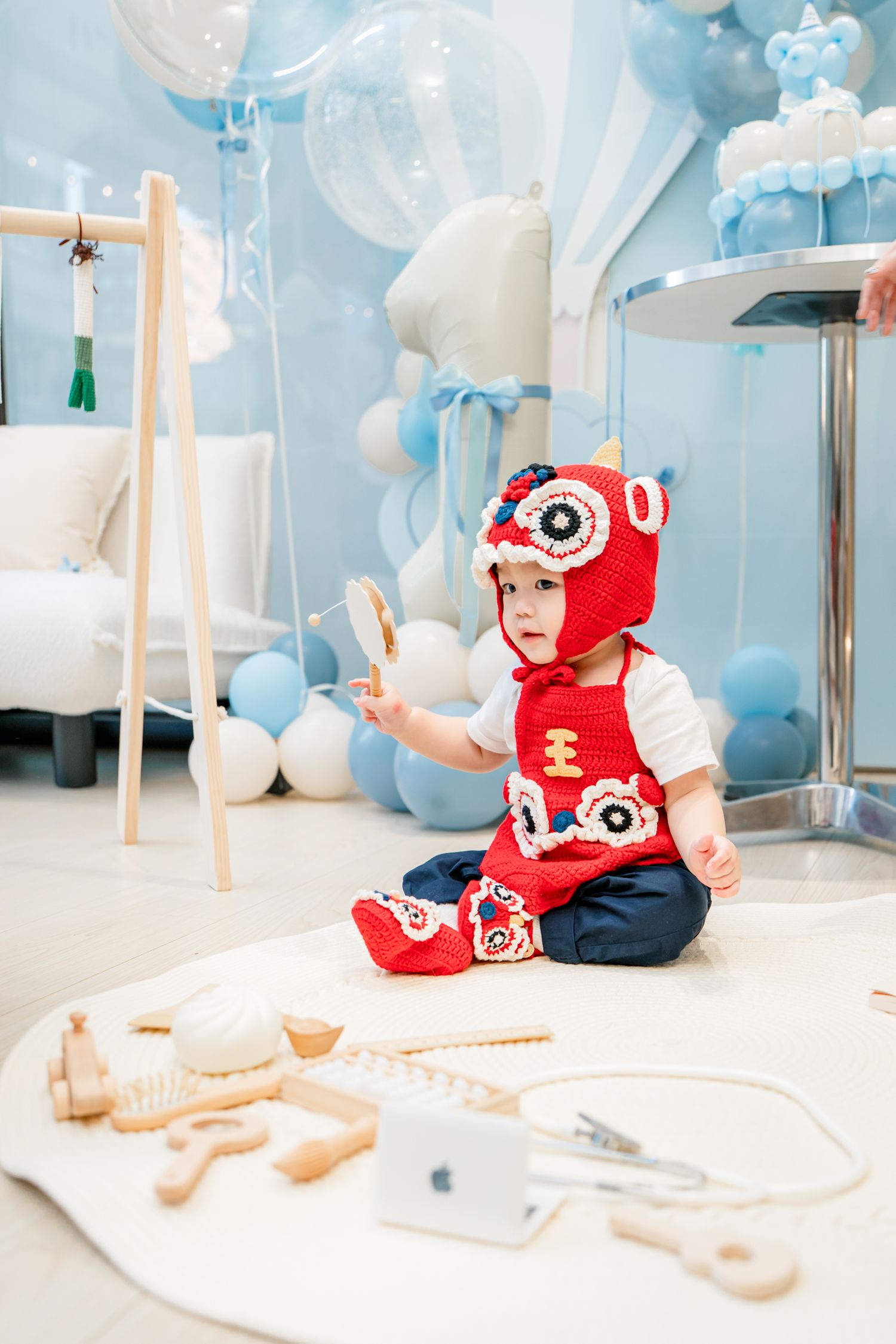 A baby in a red knit cap explores wooden toys on a white surface with blue balloons in the background.