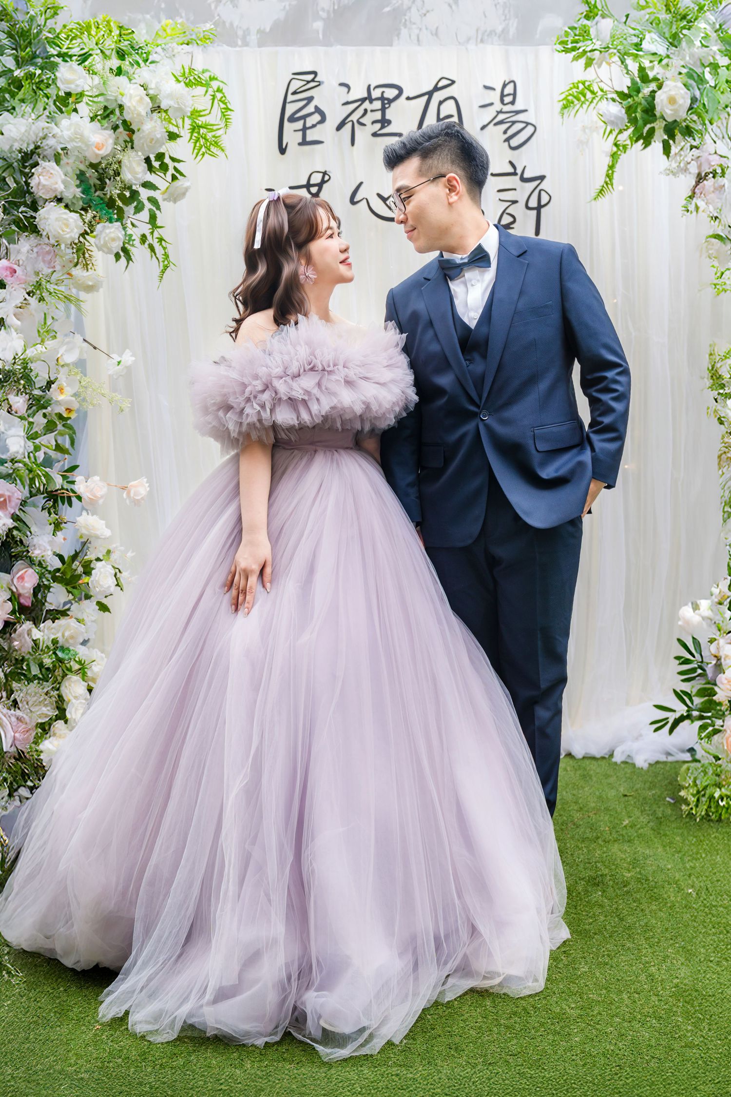 Couple poses at wedding ceremony backdrop with white floral arch decor and Chinese characters above.