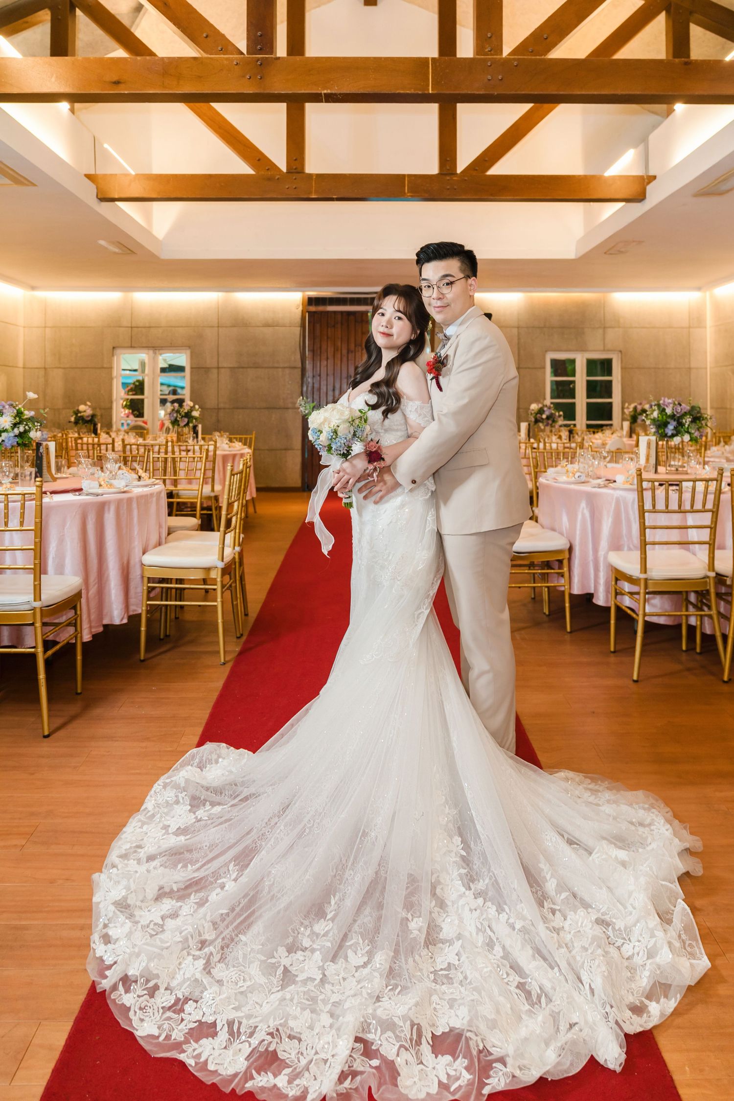 Elegant wedding reception venue features exposed wooden beams, pink table settings and gold chairs with a red carpet aisle.
