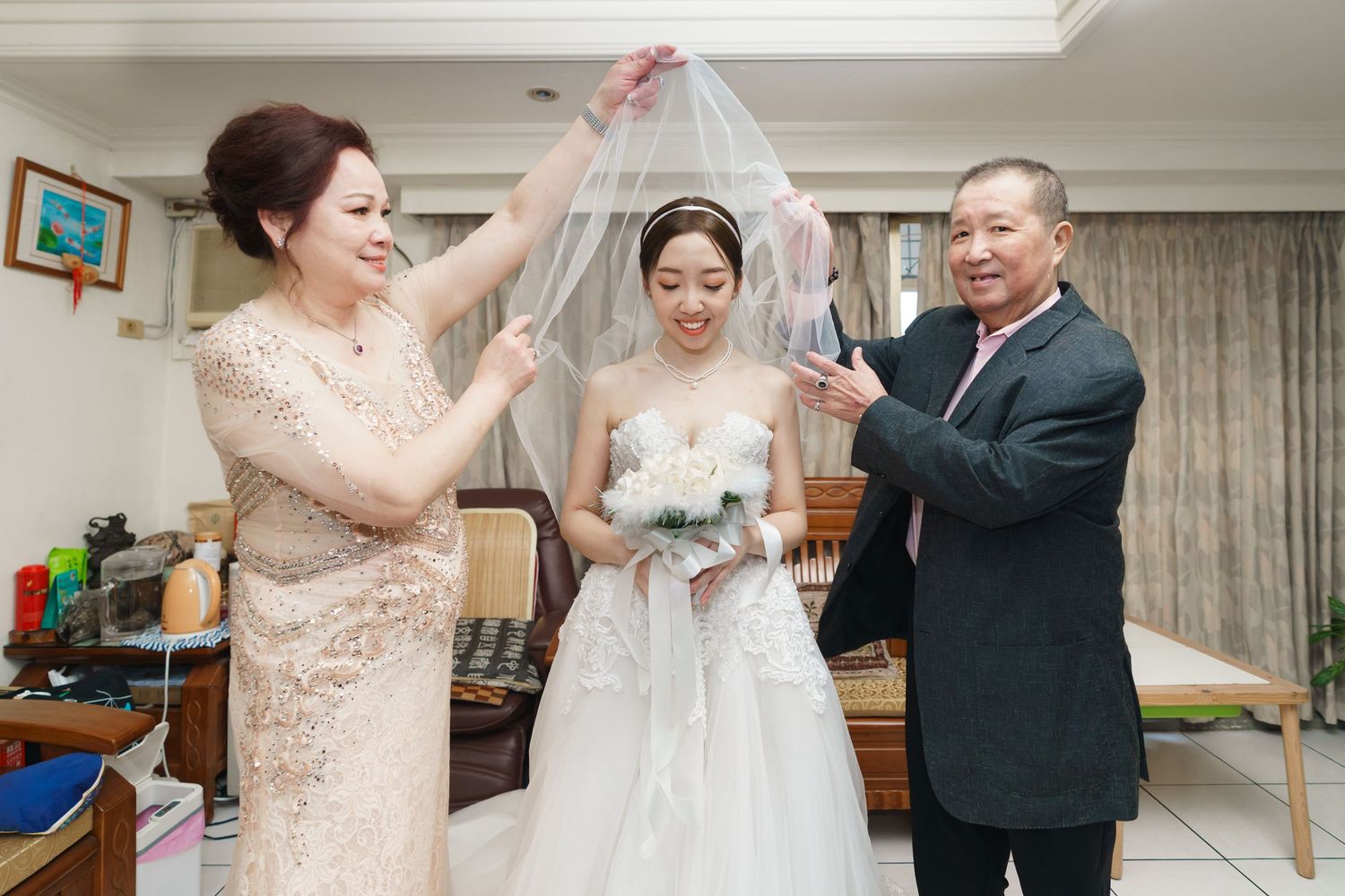 A wedding celebration scene with family members adjusting a beautiful white veil over the bride in her gown.