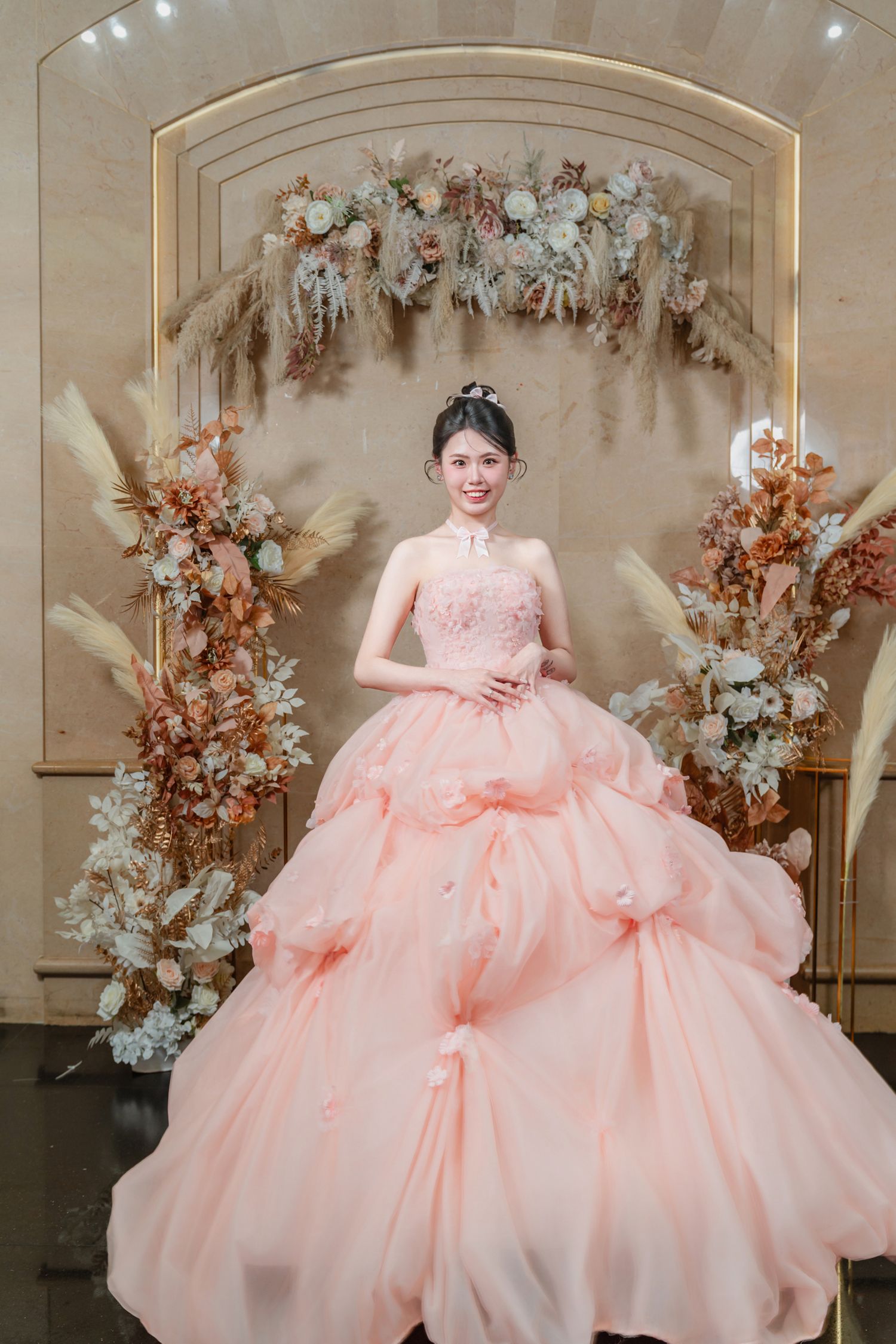 A stunning peach pink ballgown with layered tulle skirt in front of floral arch decor at an elegant photoshoot setting.