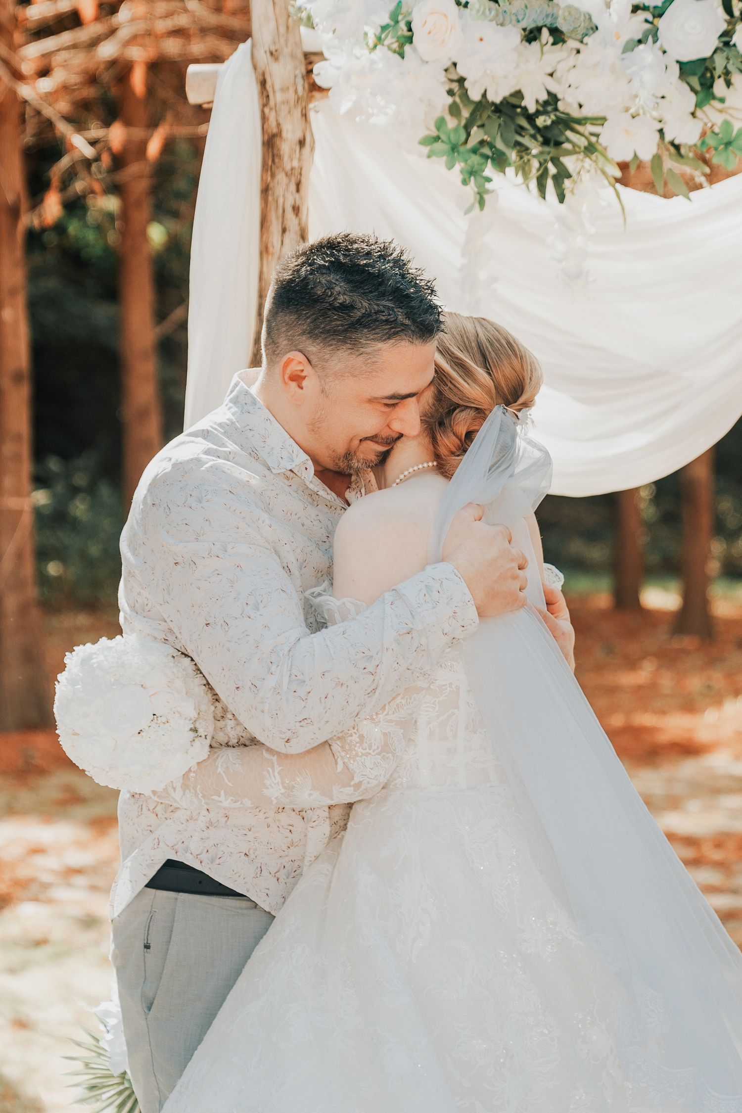 A tender moment captured at an outdoor wedding ceremony with white decor and greenery in a rustic woodland setting.