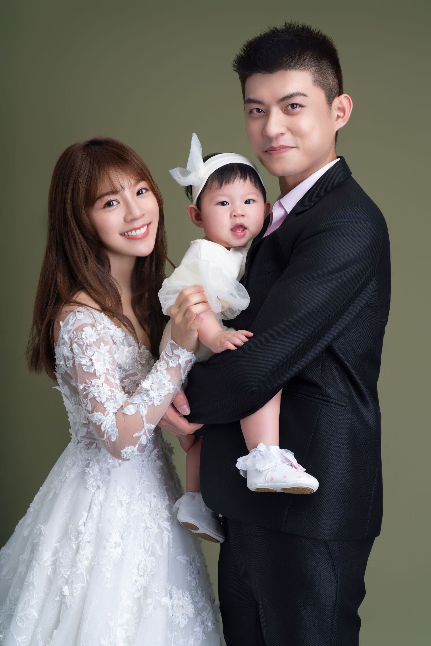 A family portrait featuring adults in wedding attire and a baby in white against a green background.
