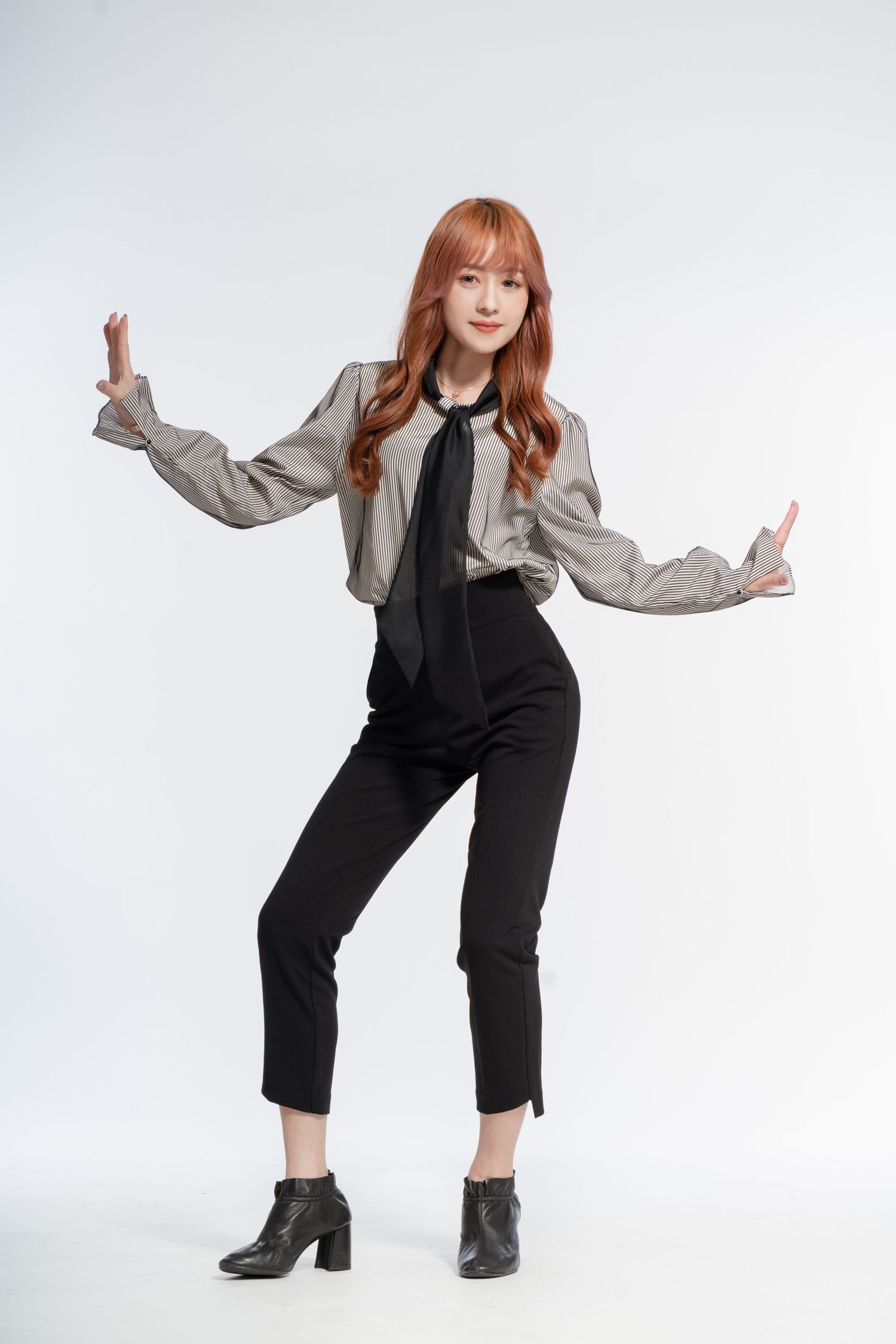 Person wearing gray cropped jacket and black jumpsuit strikes playful pose against white background.