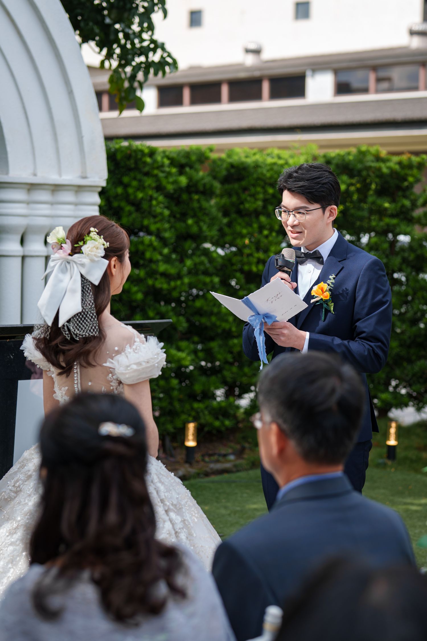 A couple exchanges vows during an outdoor wedding ceremony under a white gazebo while guests look on.