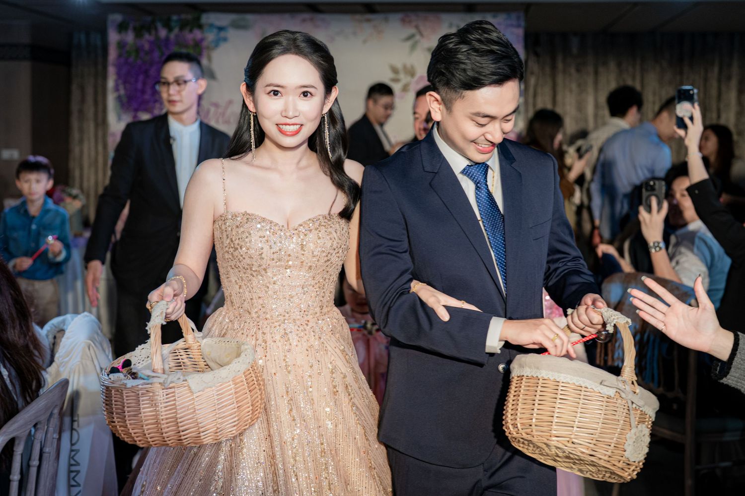 A couple in formal attire carries woven baskets at an elegant event, with the woman wearing a sparkly gold dress.