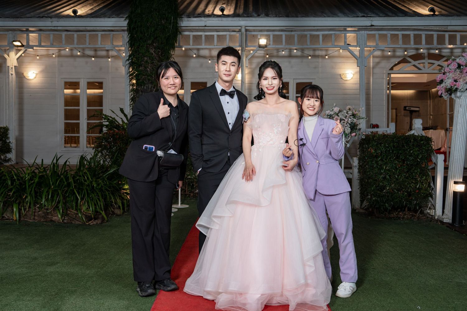 People in formal attire pose together at an elegant wedding reception venue with decorative lighting.