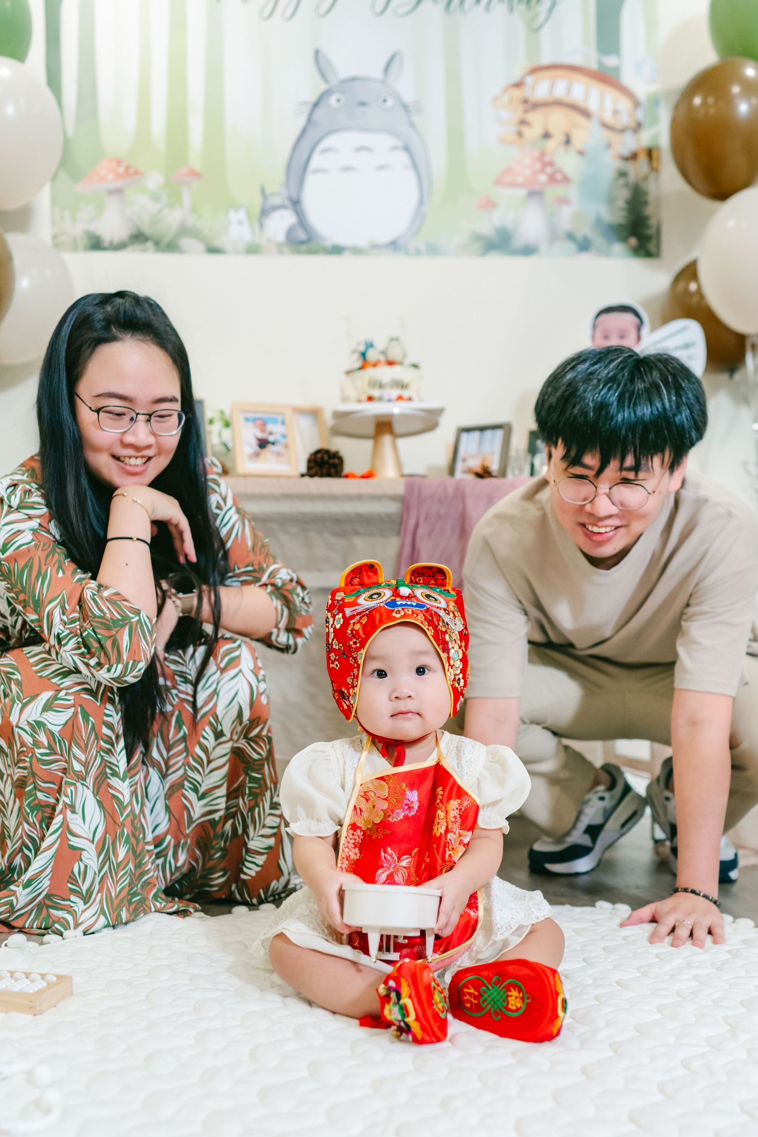 Baby wearing traditional red Chinese New Year outfit sits on floor with Totoro decorations in background.