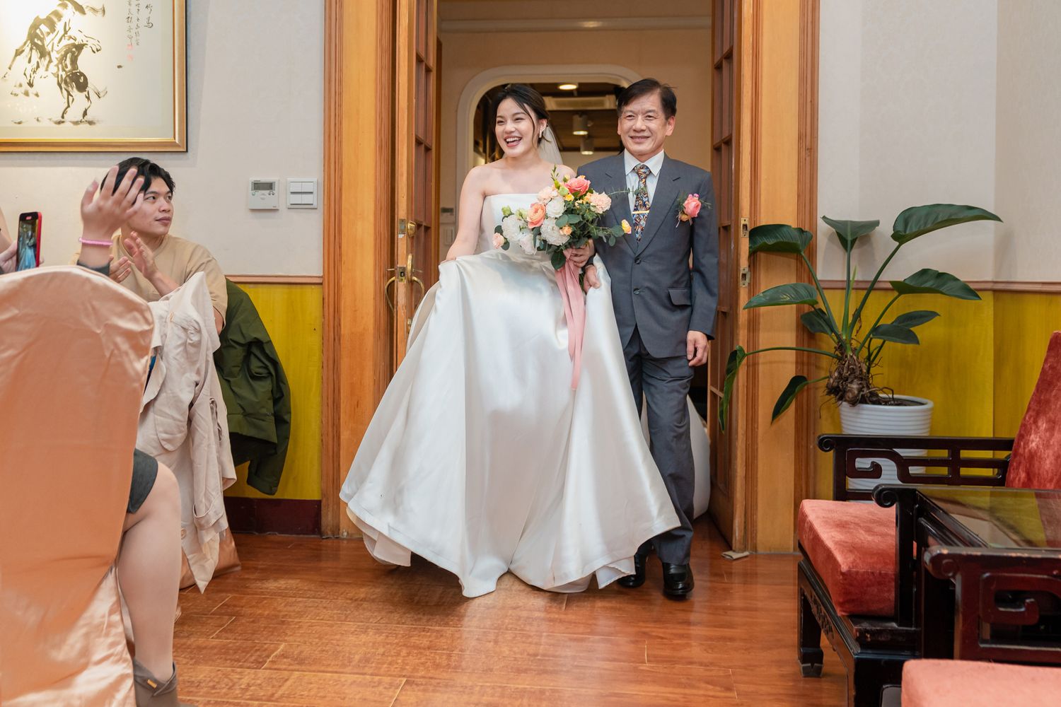 A bride in a white satin gown enters through wooden doors with a companion during a traditional wedding ceremony.