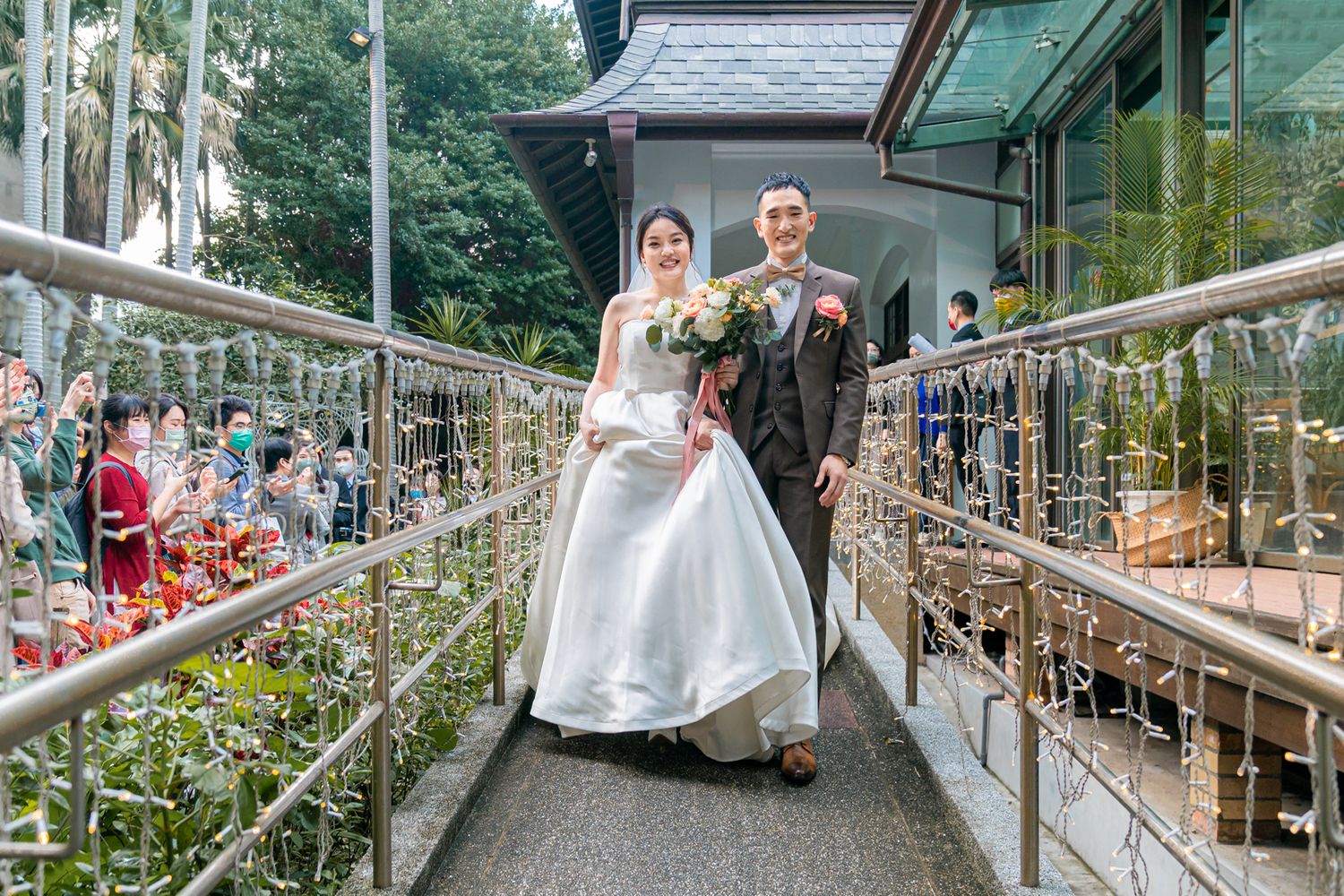 A bride and groom stand on a bridge walkway with guests watching from below at a wedding ceremony.