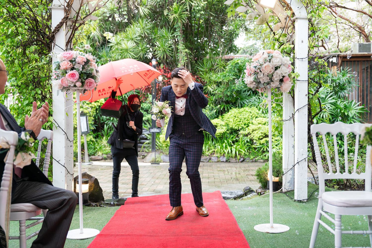 A celebratory moment on a red carpet walkway beneath a floral arch with decorative umbrellas in a garden setting.