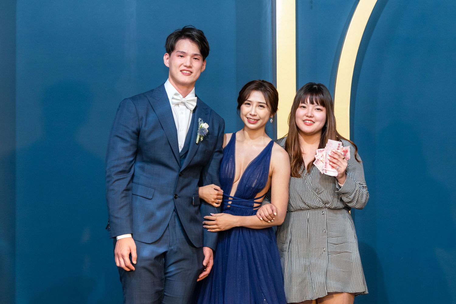 Three friends in formal attire pose together against a blue wall at an elegant event.