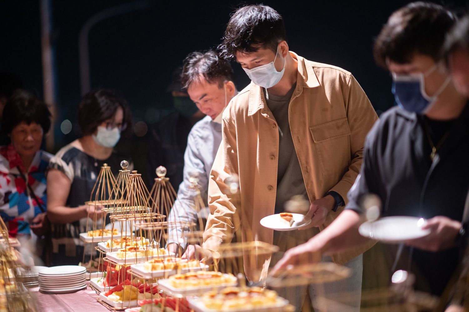 People wearing face masks gather around a buffet table serving various dishes at an indoor event.