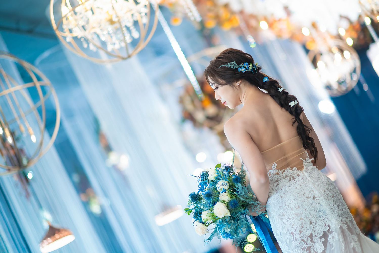 A backlit silhouette of a bride in a white lace gown holds a blue bouquet beneath crystal chandeliers.