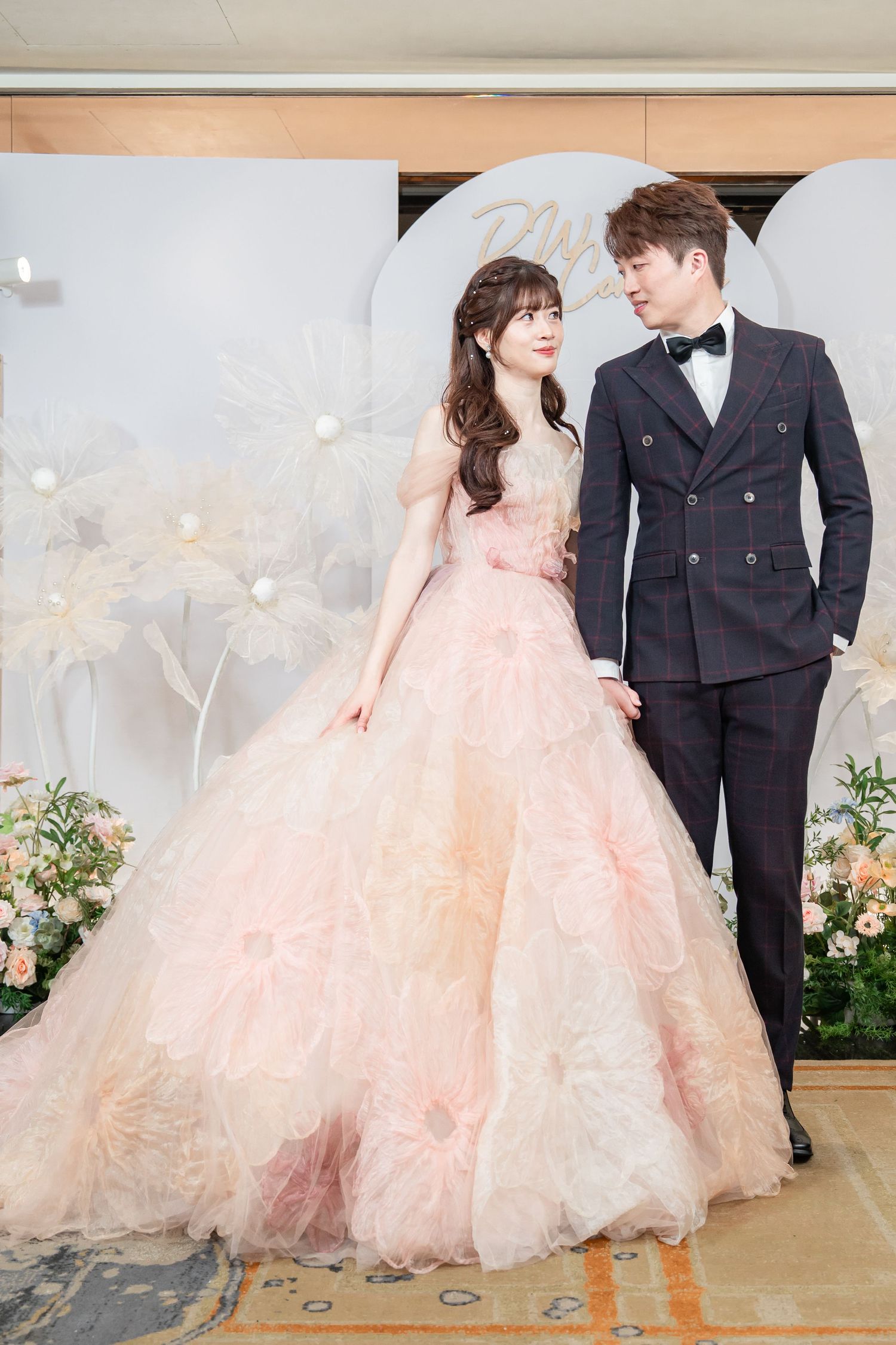 Couple posing at wedding reception, woman in pink ballgown, man in dark suit with bow tie, floral backdrop.