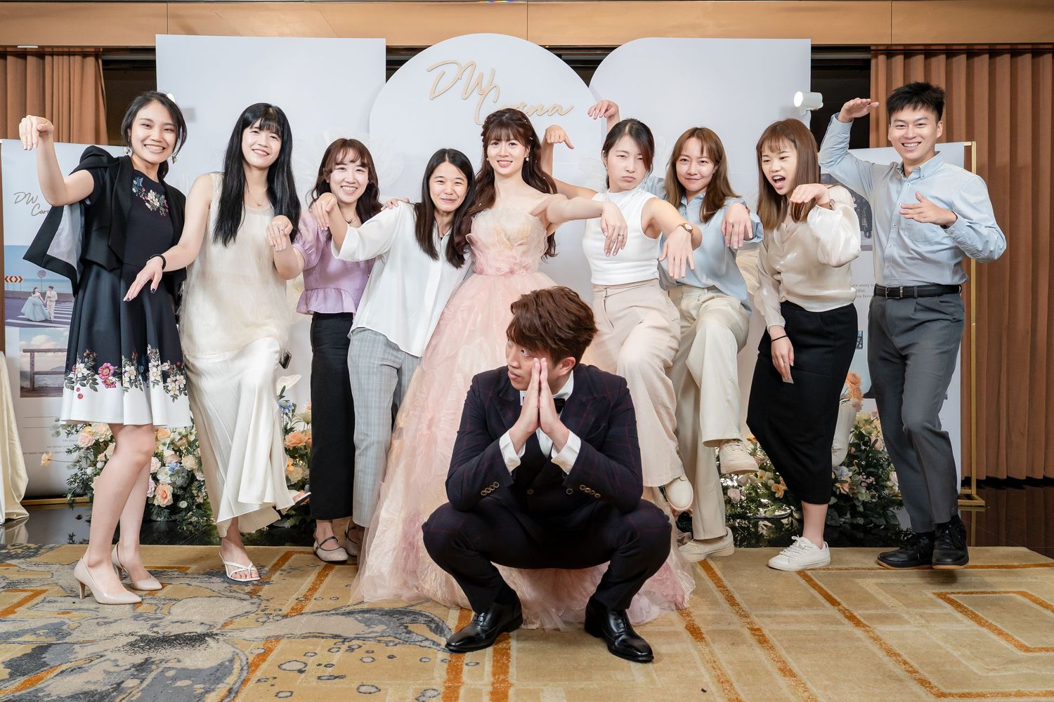 Group of friends posing joyfully at a wedding reception, with groom squatting in front and bride in pink gown at center.