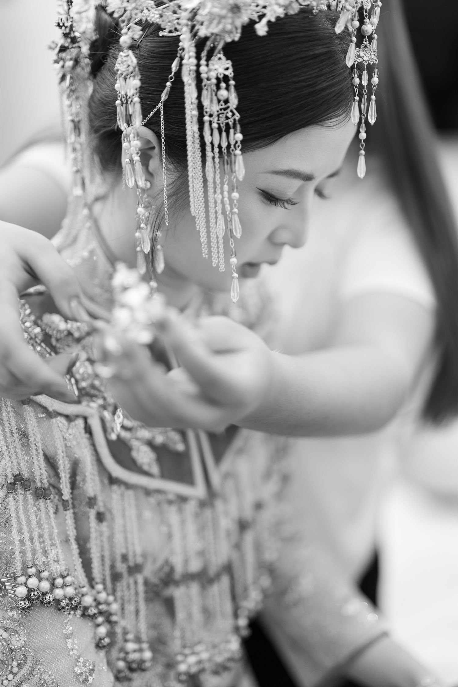 Black and white photo of a bride in an ornate beaded Chinese wedding headdress and embroidered gown as helping hands adjust her attire.