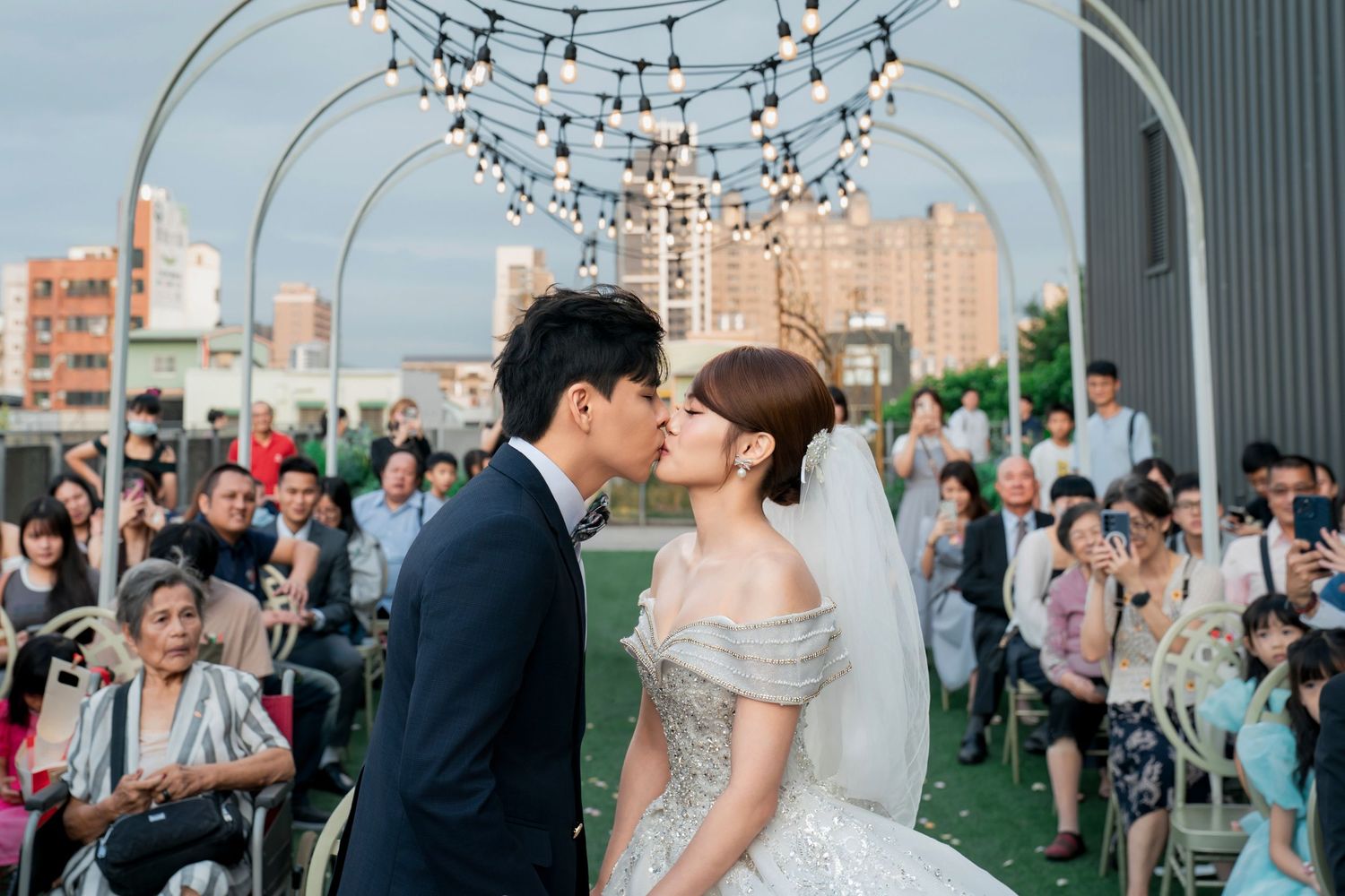 A couple shares a romantic kiss at an outdoor rooftop wedding ceremony with string lights and guests seated behind them.