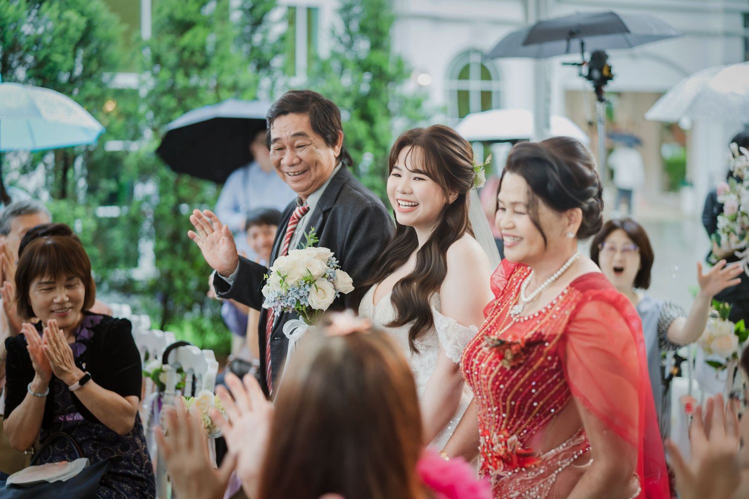 Joyful wedding guests celebrate under umbrellas at an outdoor garden ceremony with white floral decorations.