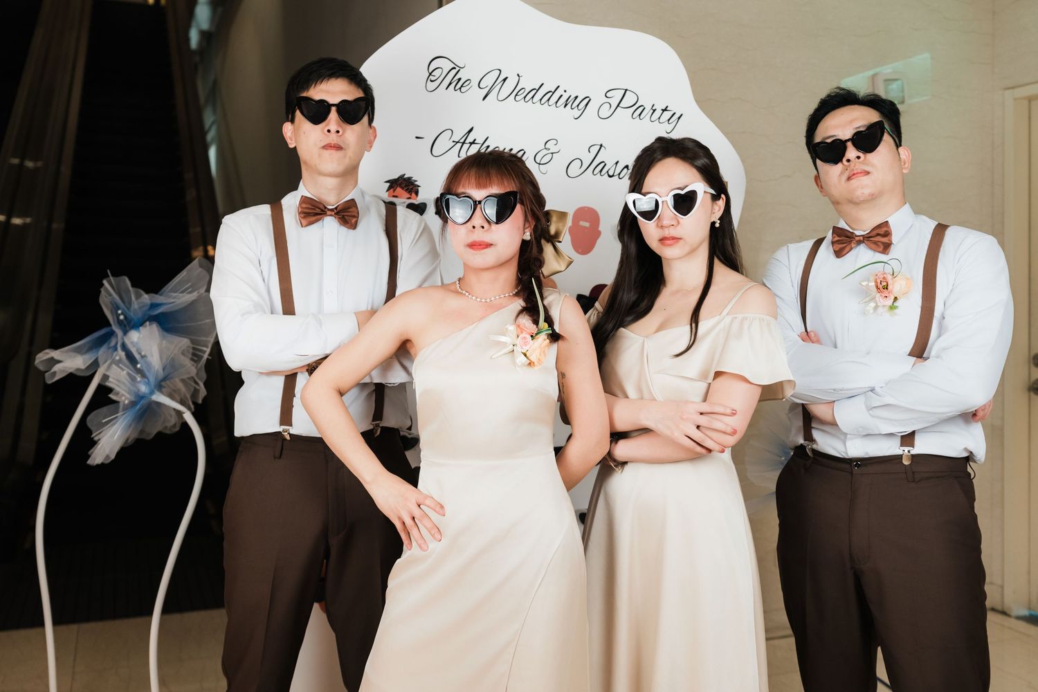 Wedding party members pose together wearing matching brown suspenders and sunglasses at indoor celebration.