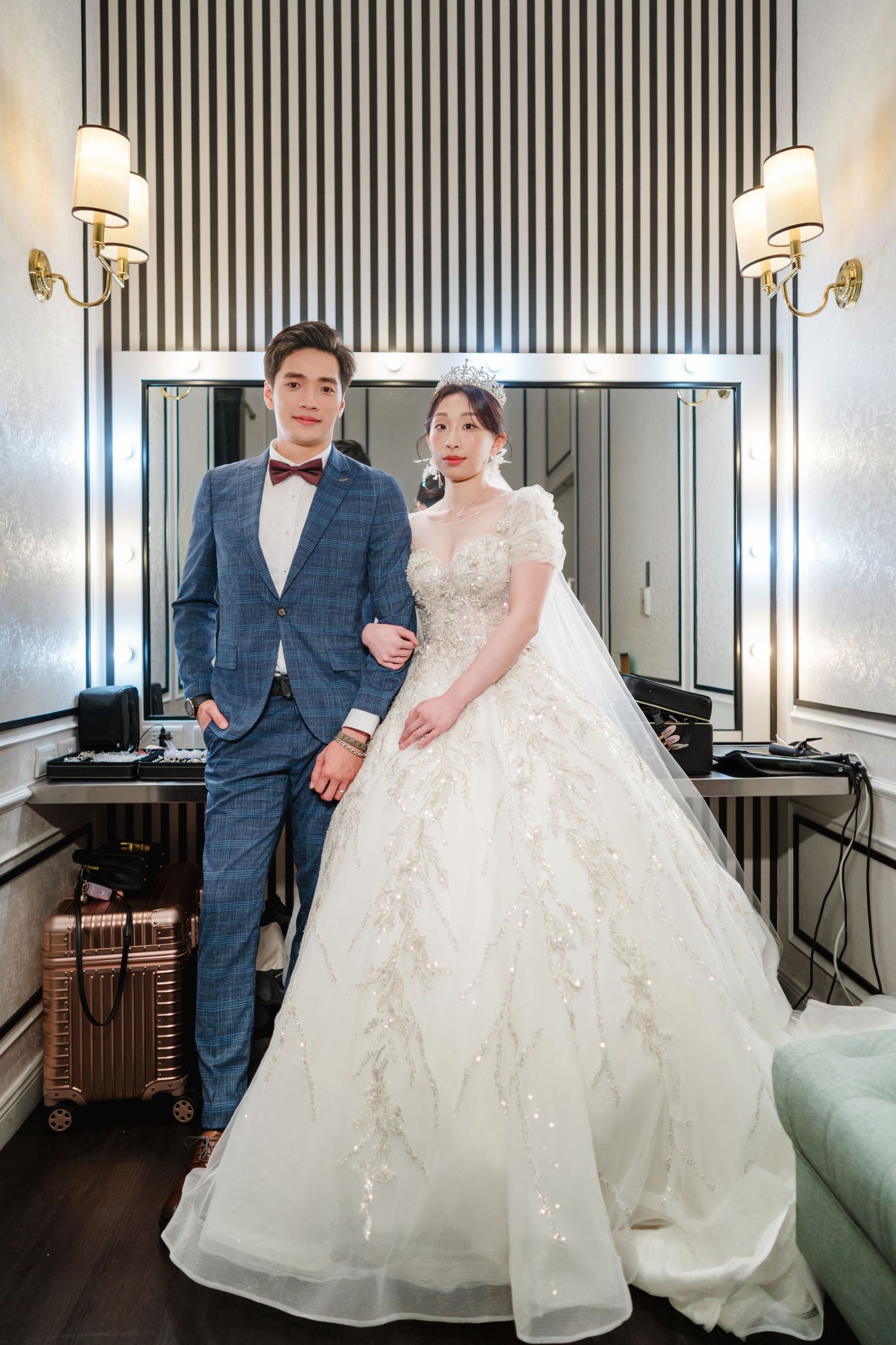 A couple poses in formal wedding attire with a black and white striped wall and wall sconces in the background.