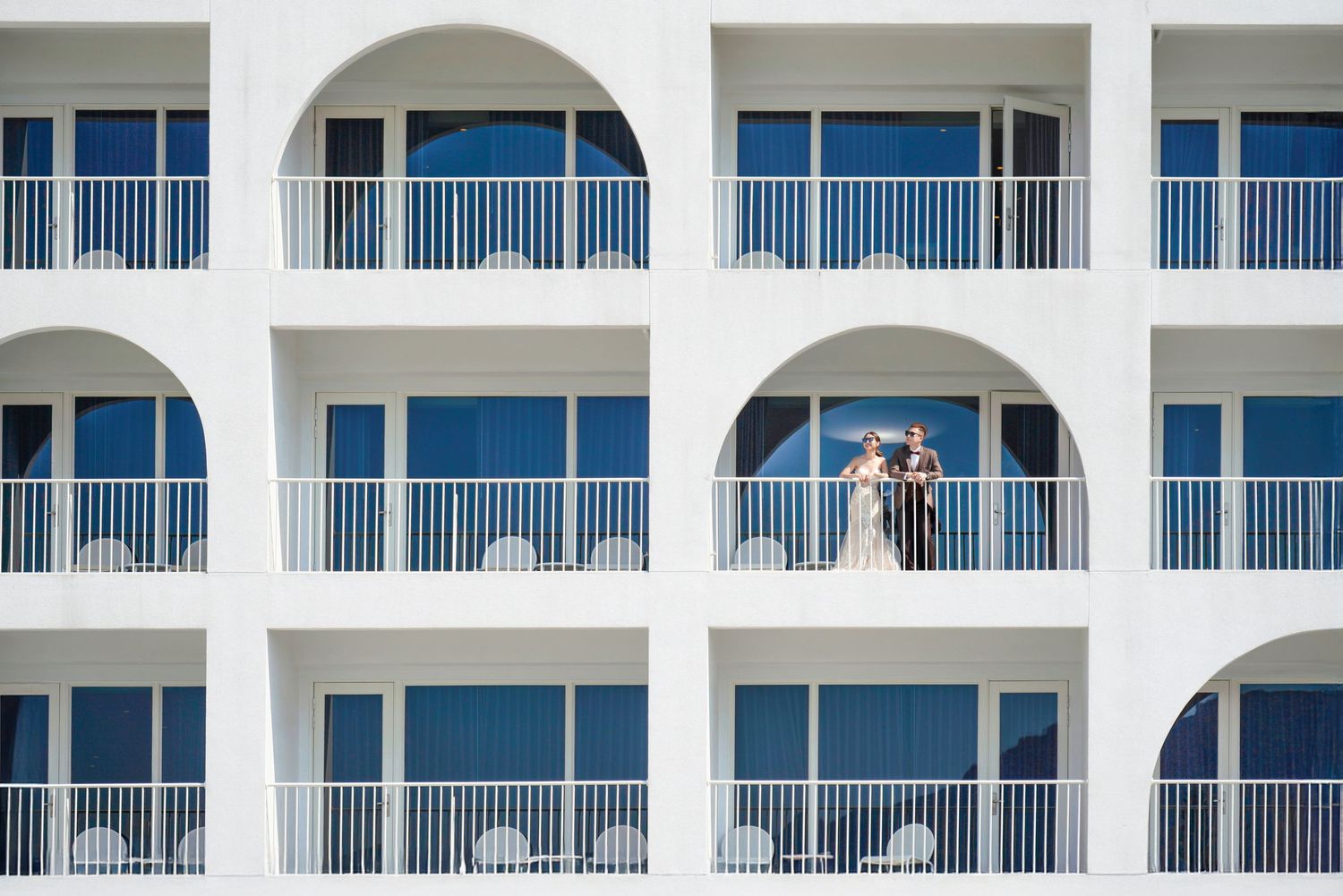 A white hotel building exterior with arched balconies overlooking the ocean, featuring white railings and blue windows.