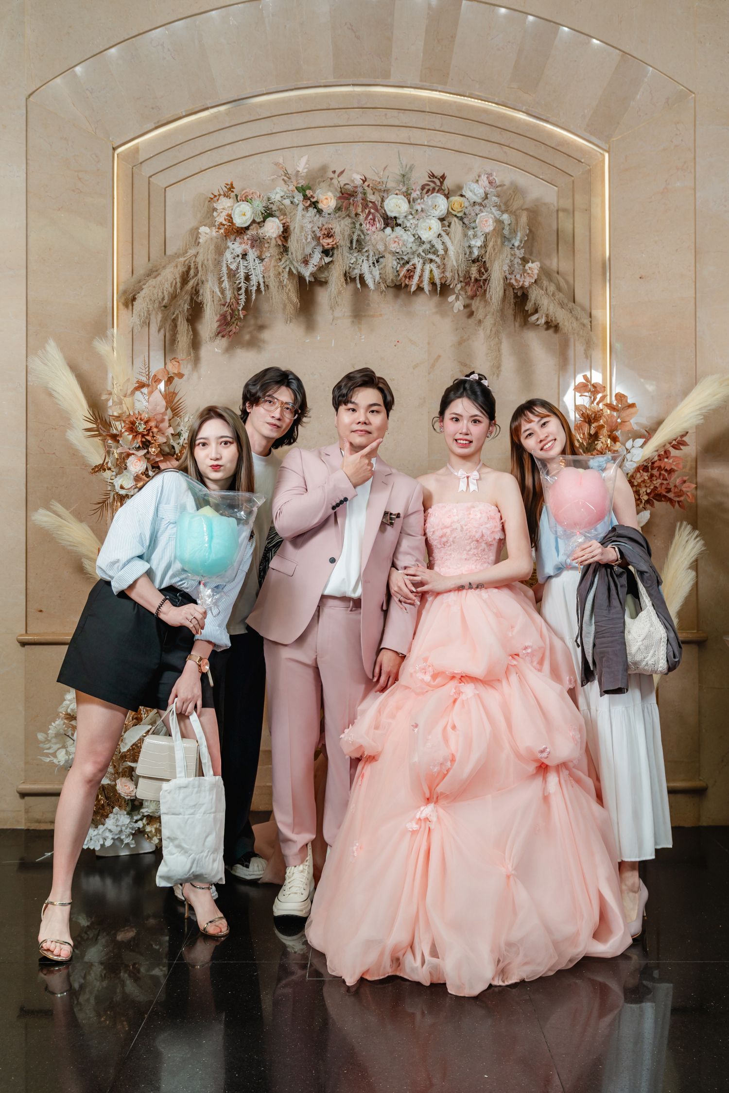 Group photo against floral arch backdrop with formal attire including pink gown and coordinated pastel outfits.