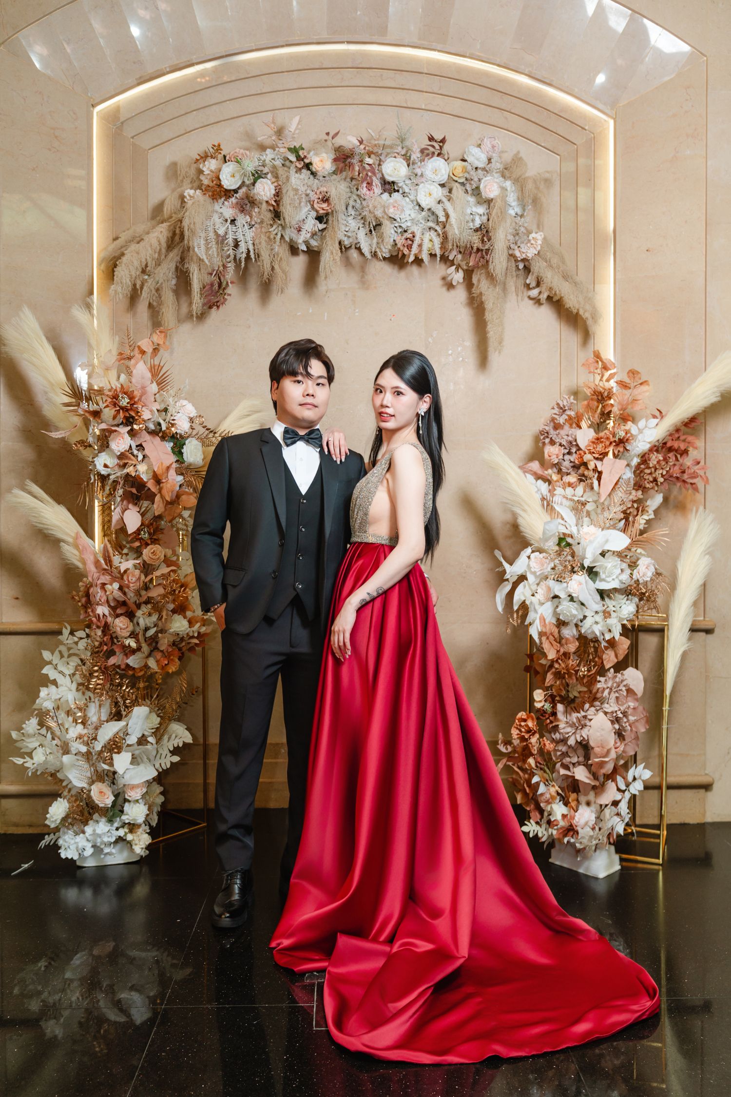 Couple poses in formal attire against an elegant floral arch backdrop with dried flowers and cream decor.
