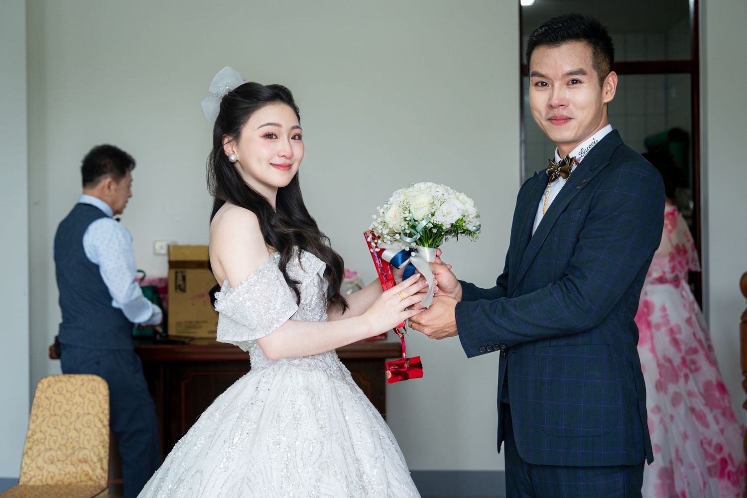 Bride in white lace gown and groom in black suit exchanging bouquet of white roses at wedding ceremony.