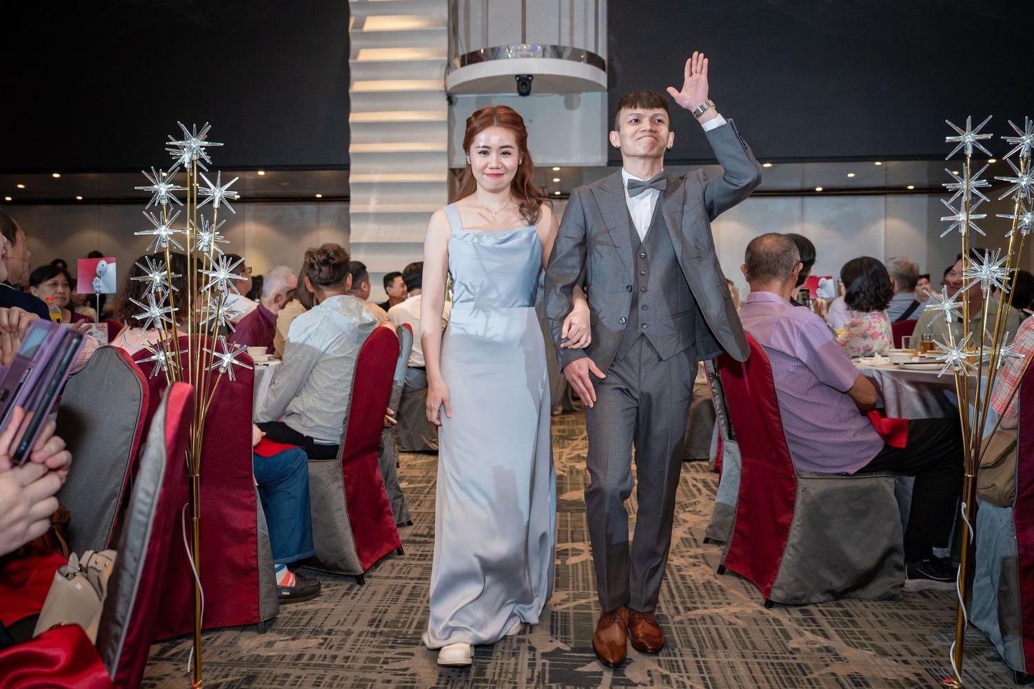 A bride and groom walk hand-in-hand through a decorated wedding banquet hall, smiling joyfully at their reception.