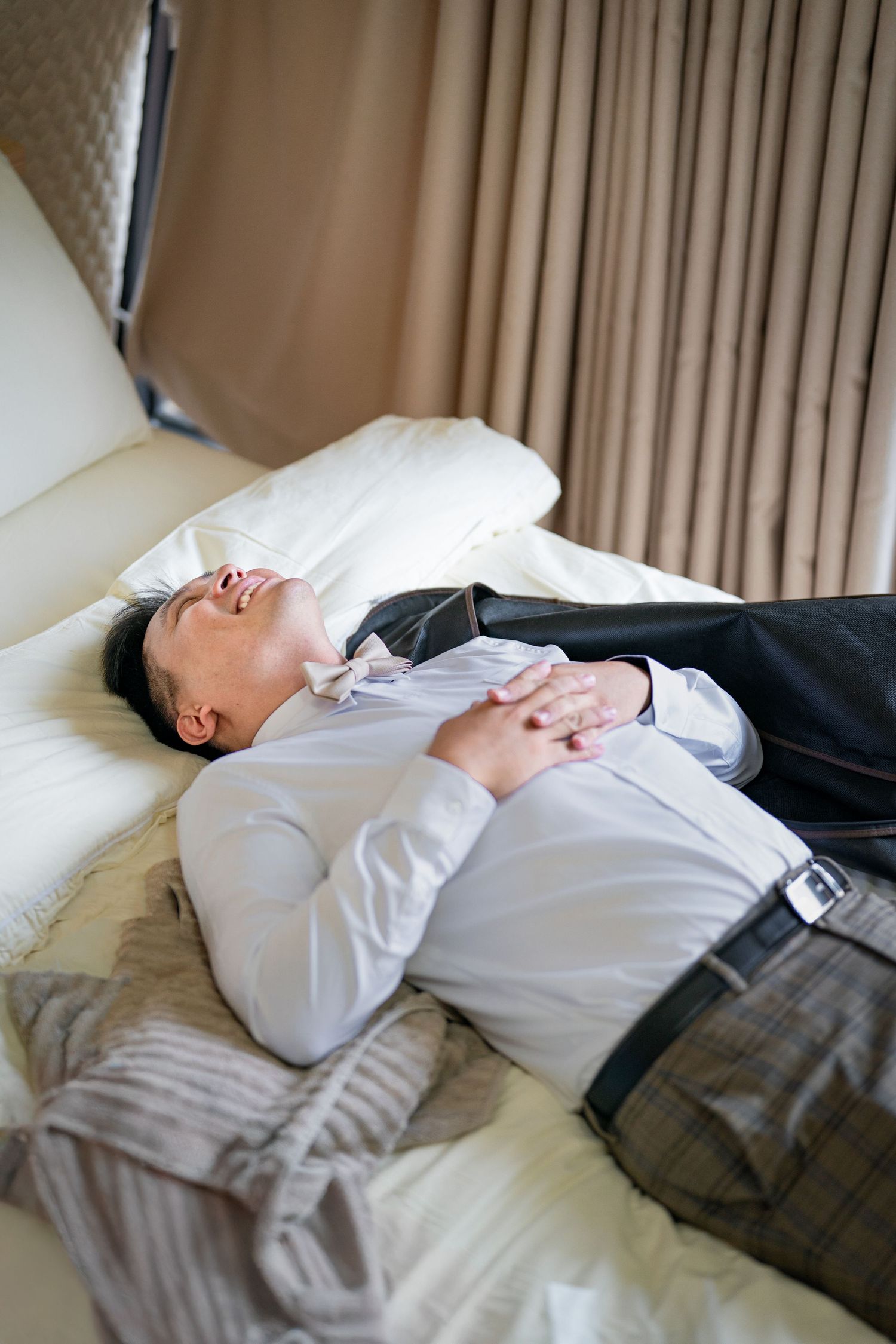 A person in business attire lies exhausted on a white hotel bed with curtains in the background.