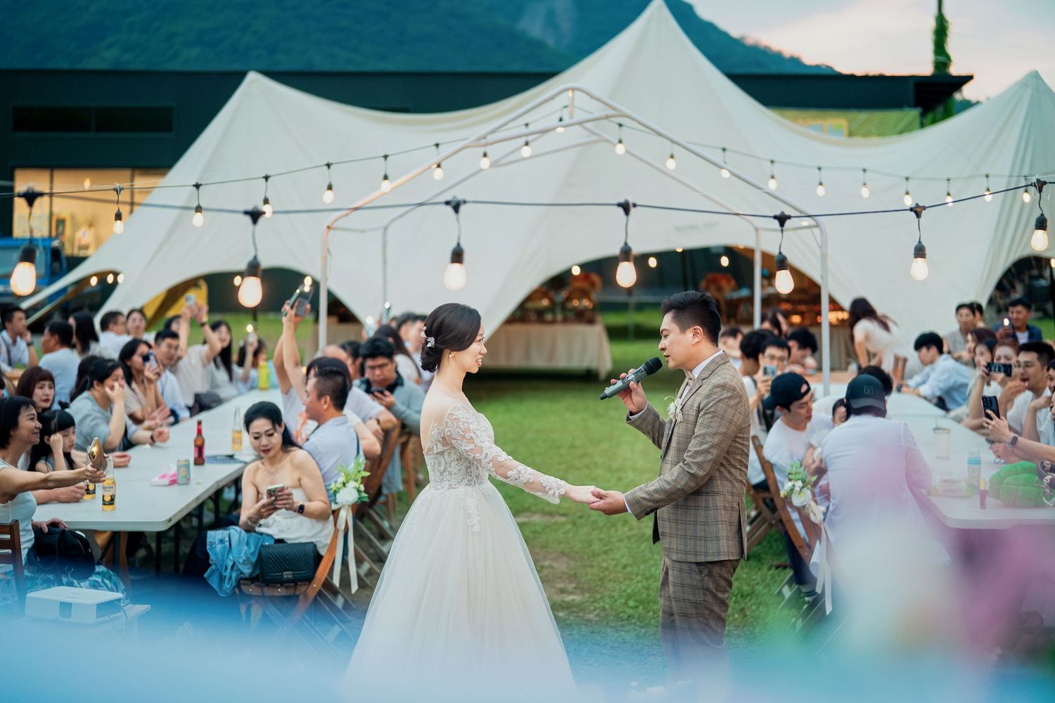 Outdoor reception under white tent canopy with string lights and guests seated at tables.