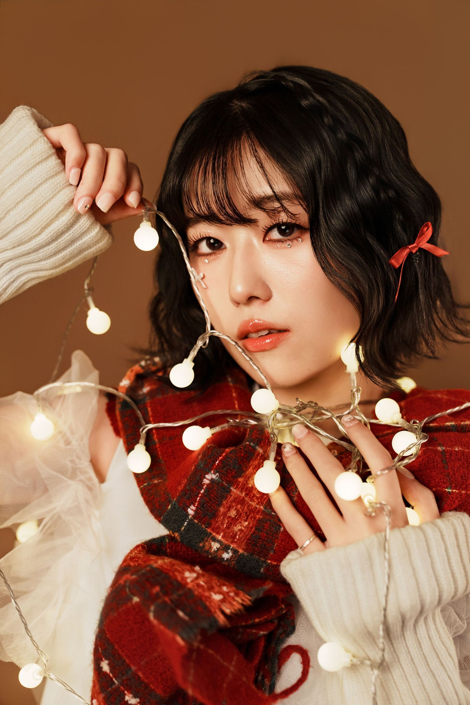 Portrait with glowing string lights against brown background wearing red plaid and cream sweater sleeves.