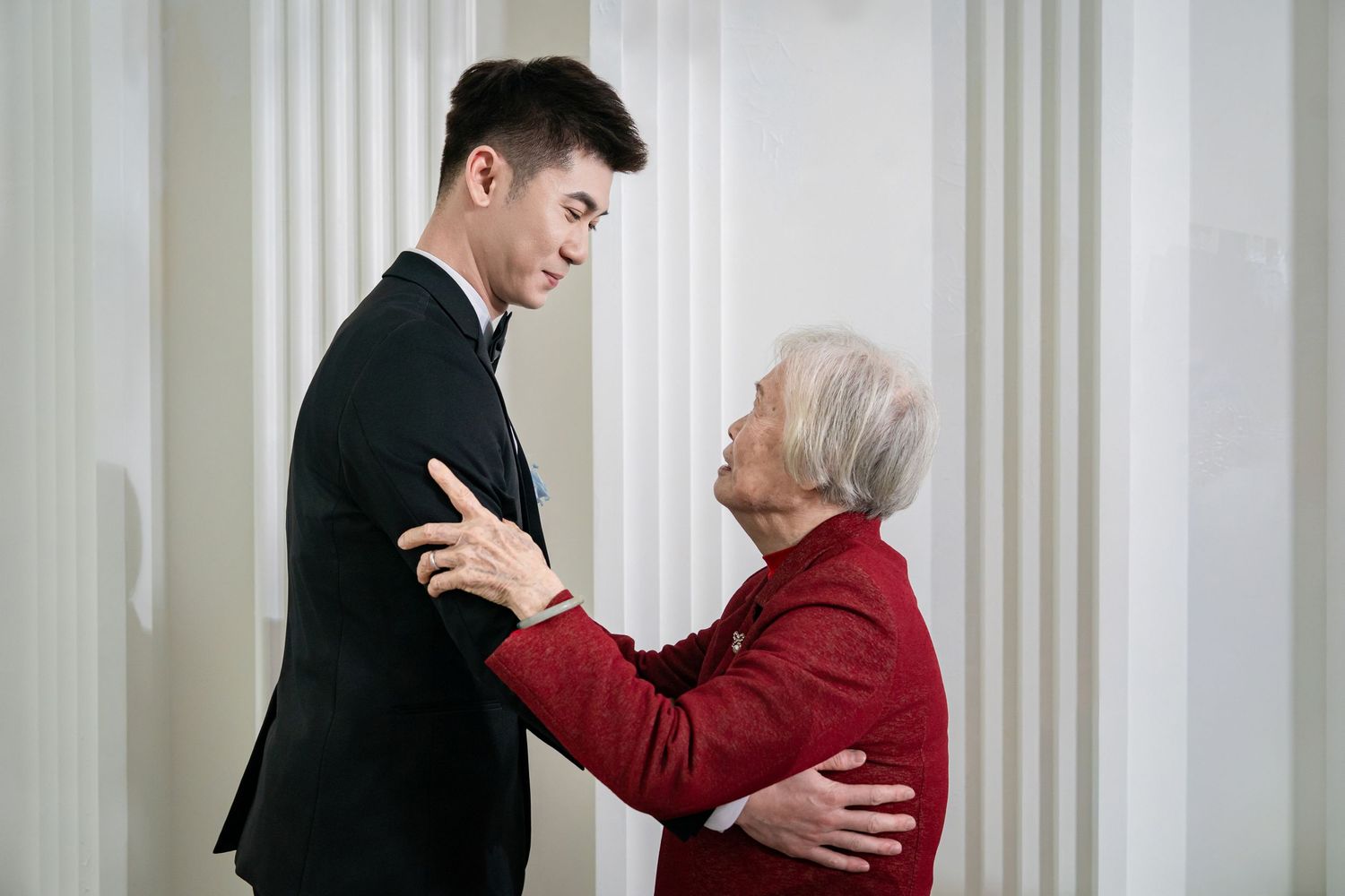 An emotional moment shared between someone in a suit and an elderly person wearing red in an elegant setting.